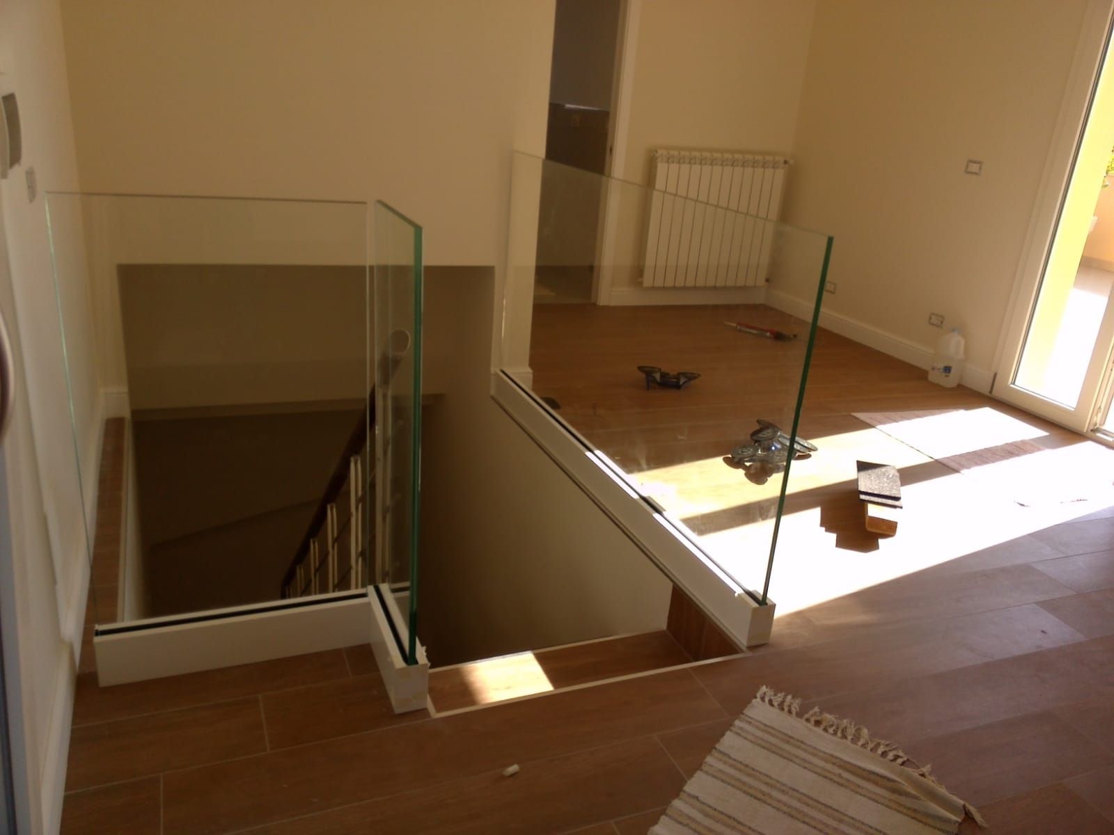 Vista interna di una scala con ringhiere in vetro e pavimento in legno che scende. La luce del sole filtra all'interno.