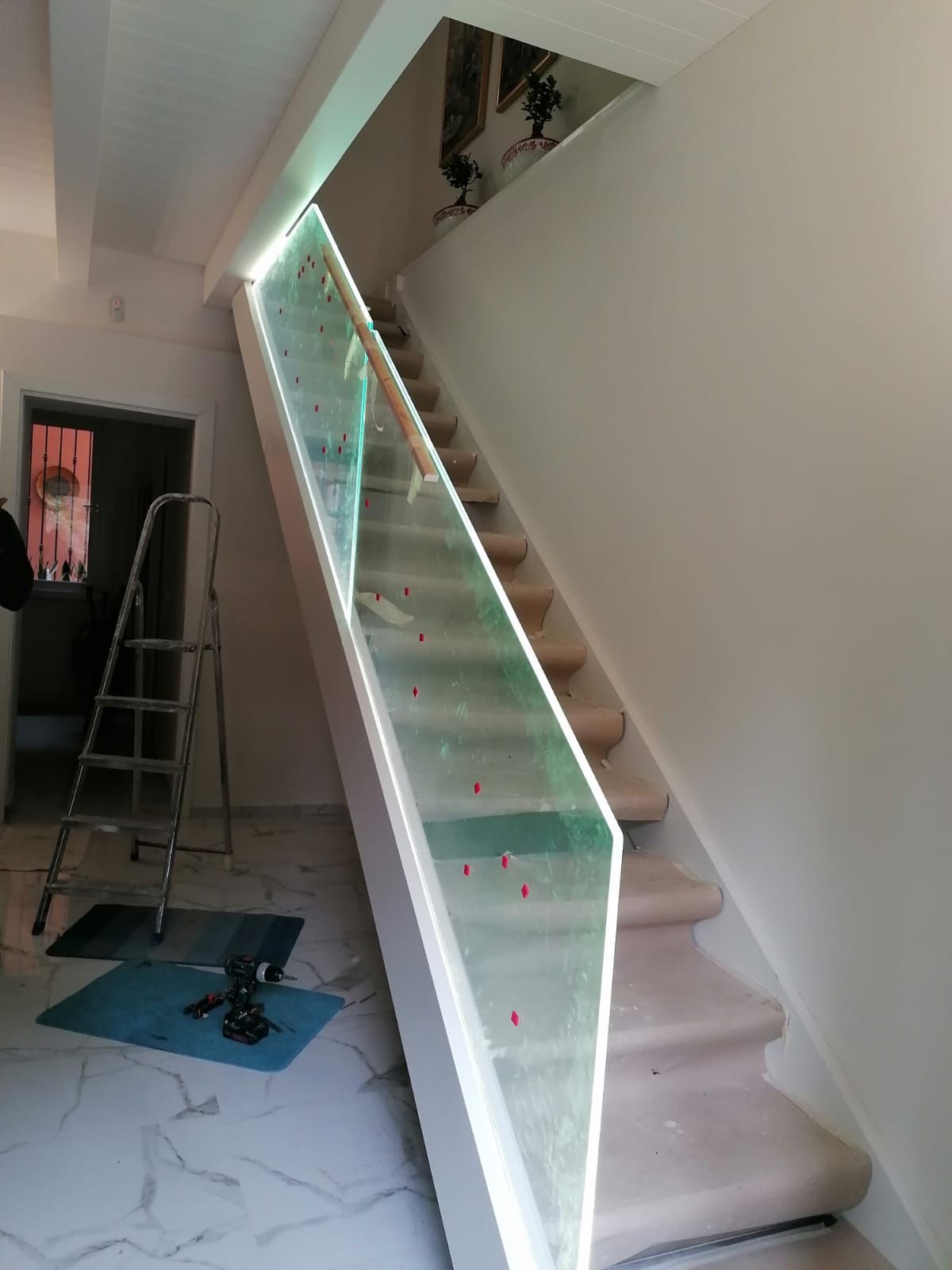 Scala con ringhiera in vetro e luci incassate, gradini rivestiti in moquette beige, pareti e finiture bianche, porta e scala parzialmente visibili.