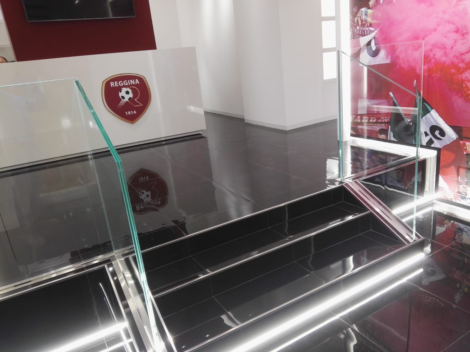Vetrina con ripiani in vetro, all'interno di un negozio, con il logo della squadra sulla parete. Piastrelle bianche e nere.