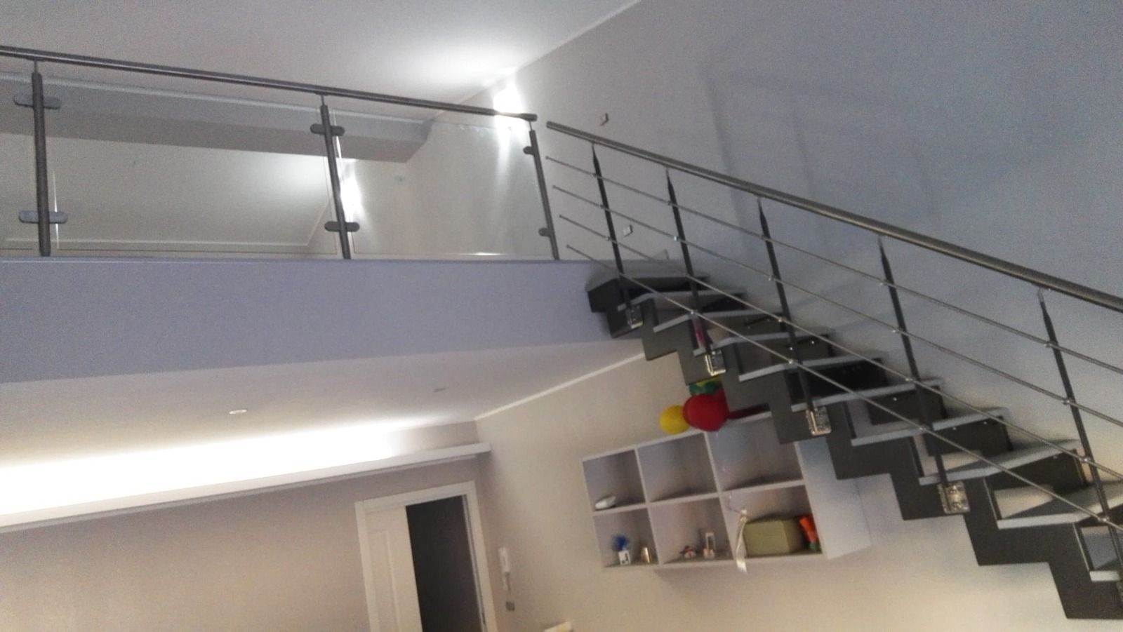 Scala con ringhiera in metallo che sale al secondo piano. Pareti bianche, gradini grigi e una sporgenza bianca.
