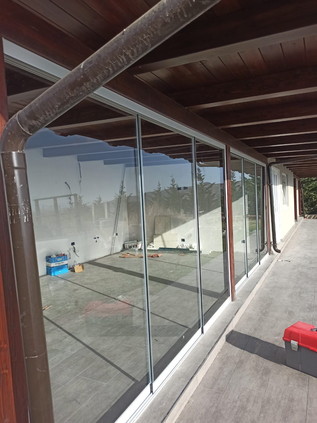 Patio con pareti in vetro, finiture in legno marrone e grondaia, che riflette l'ambiente esterno.