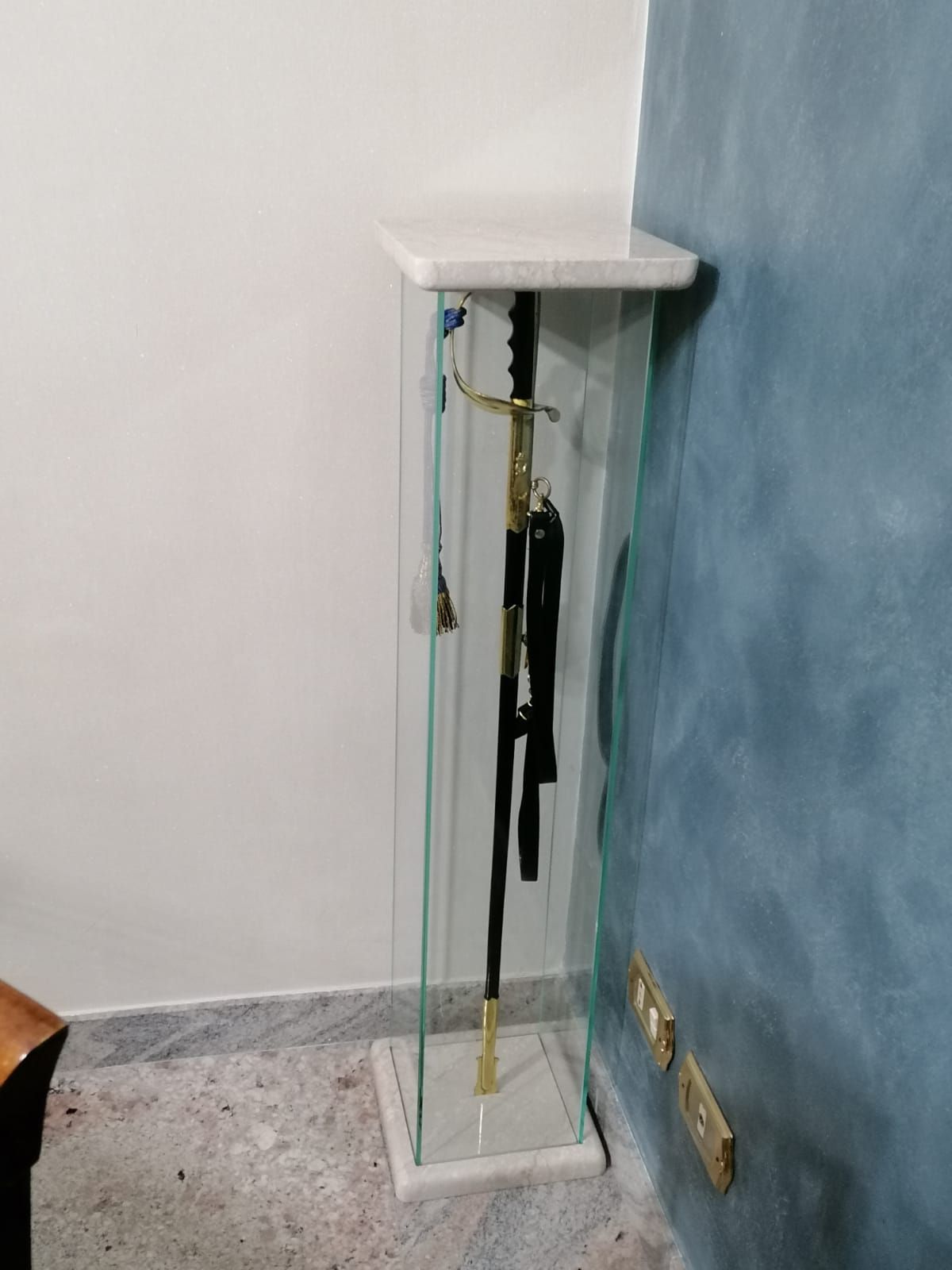 Una spada esposta in una teca di vetro con base e parte superiore in marmo, posta vicino a una parete blu.
