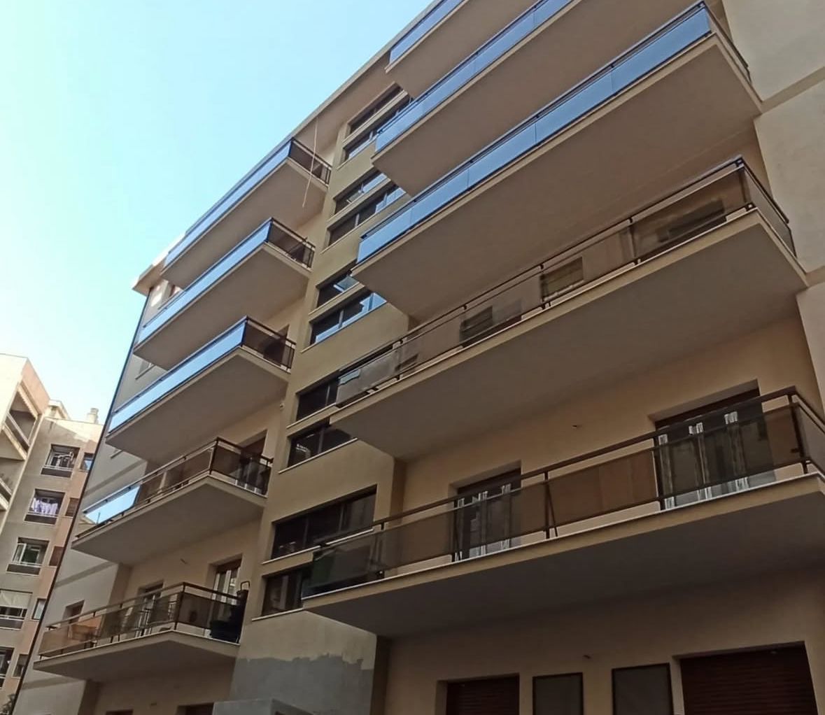 Edificio residenziale a più piani con balconi, pareti beige, ringhiere in vetro e cielo azzurro.