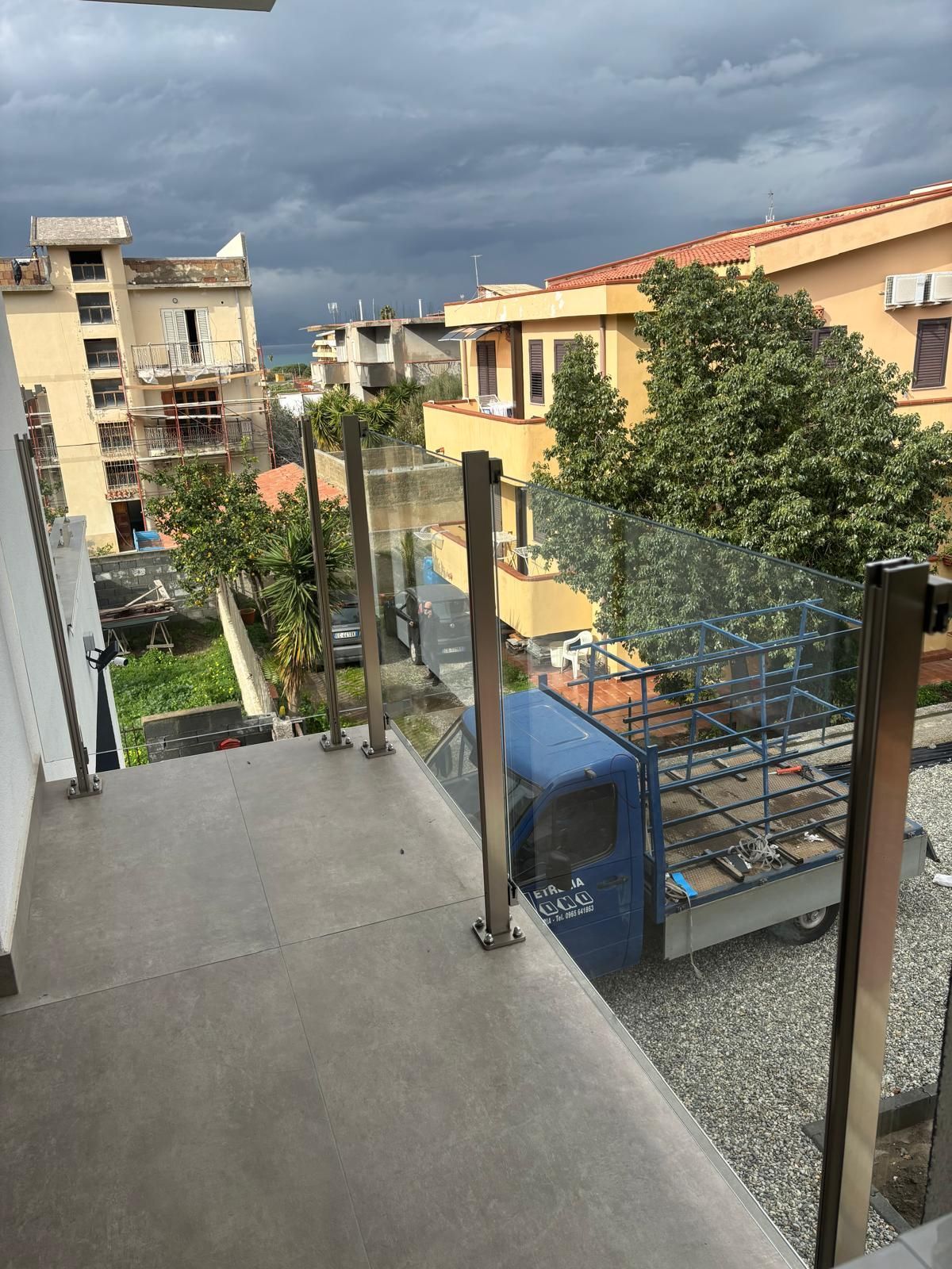 Balcone con ringhiera in vetro, con vista su edifici, alberi e un camion blu parcheggiato in una giornata nuvolosa.