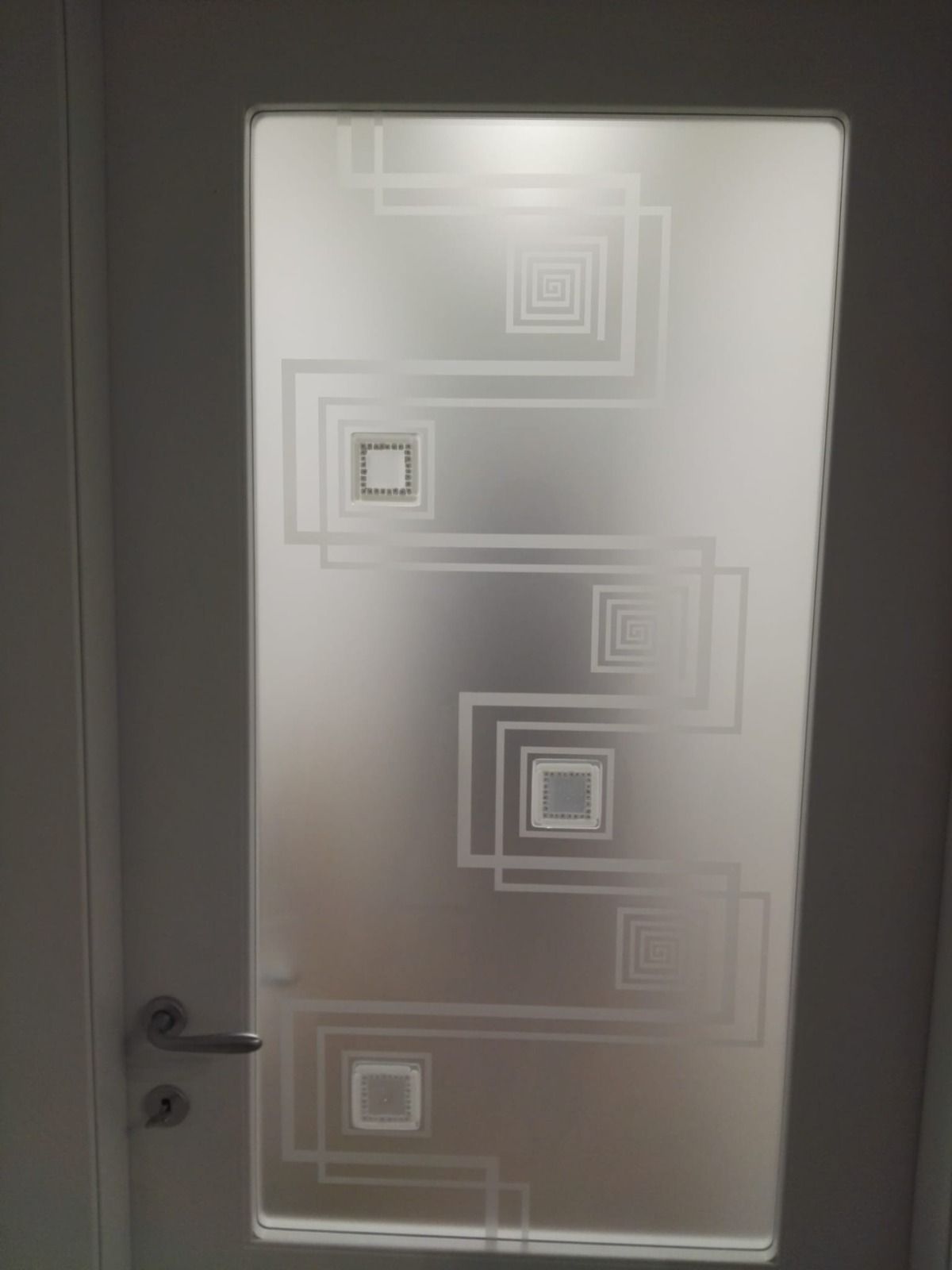 Porta bianca con pannello in vetro satinato caratterizzato da un disegno geometrico quadrato.
