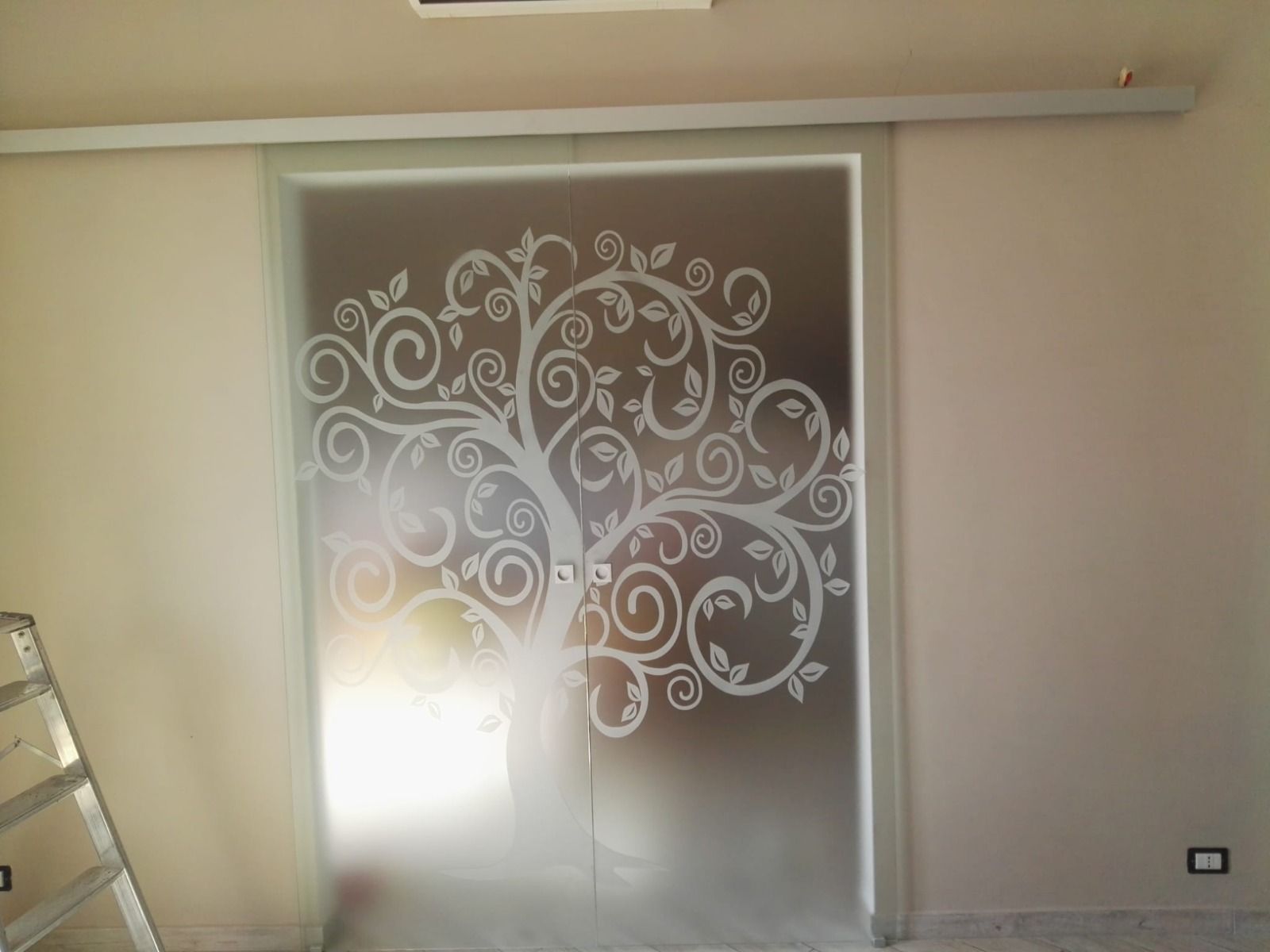 Porta scorrevole in vetro con disegno di un albero inciso, inserita in pareti beige.