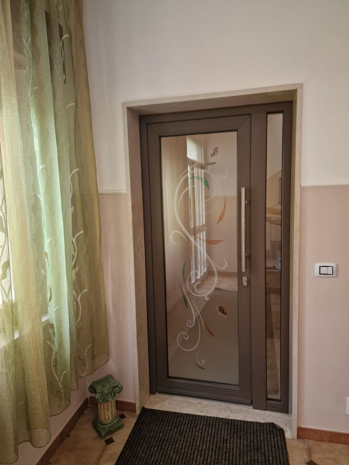 Porta d'ingresso con pannelli in vetro e motivi decorativi a spirale, accanto a una finestra laterale. Telaio marrone, tappetino scuro, pareti chiare e tenda verde.