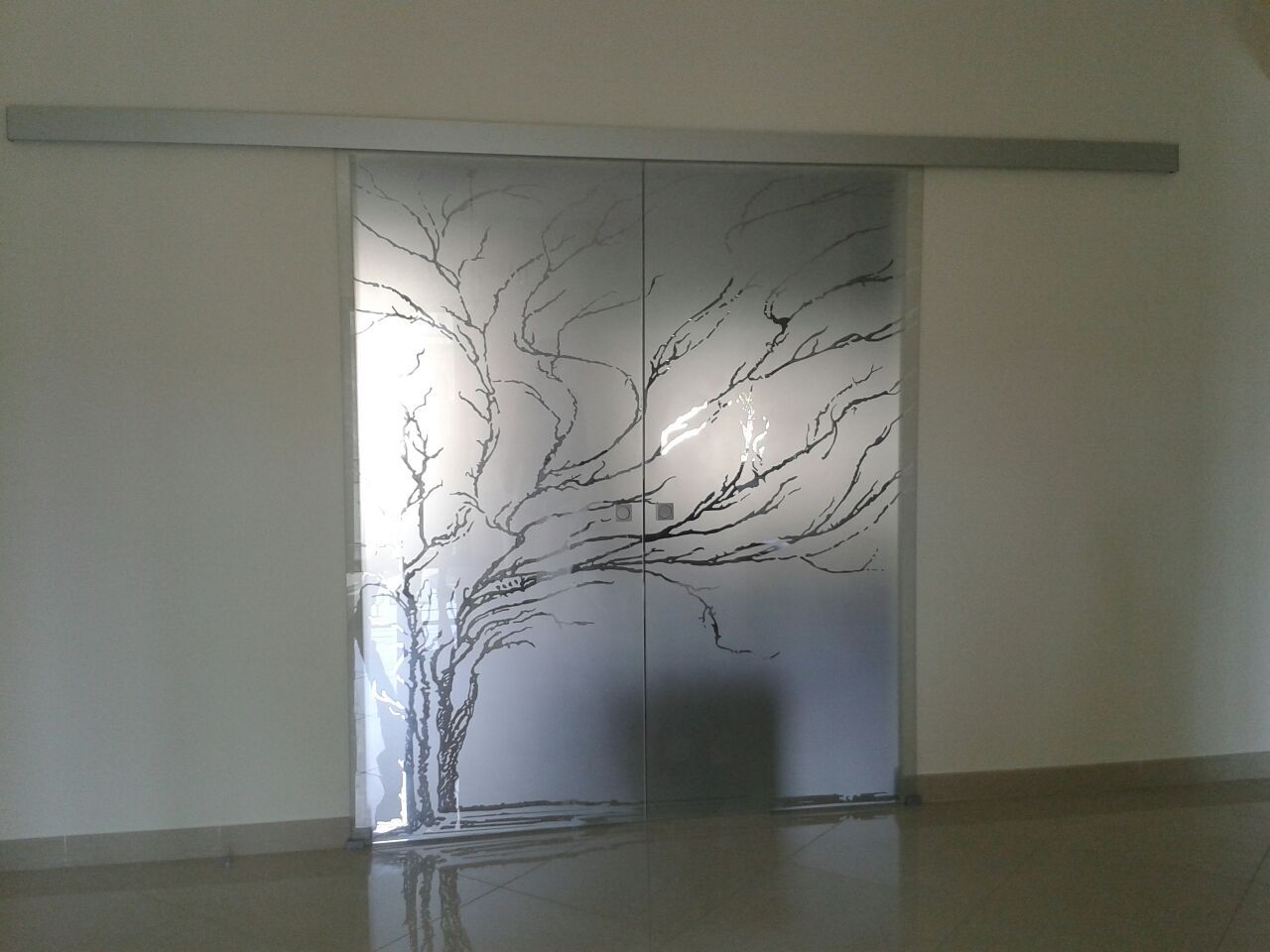 Porte scorrevoli in vetro con disegno di un albero inciso, montate su un binario argentato, inserite in una stanza dalle pareti bianche.