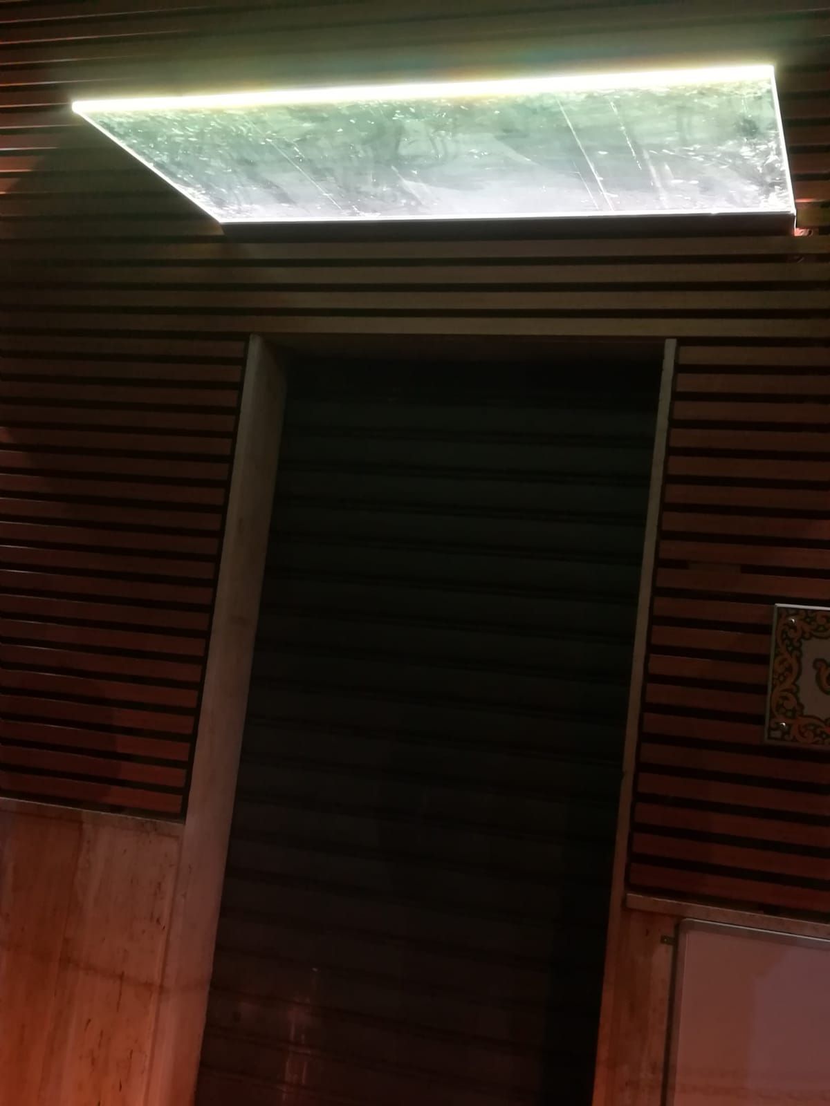 Ingresso di un edificio moderno con pannelli in legno, pensilina in vetro e illuminazione esterna.