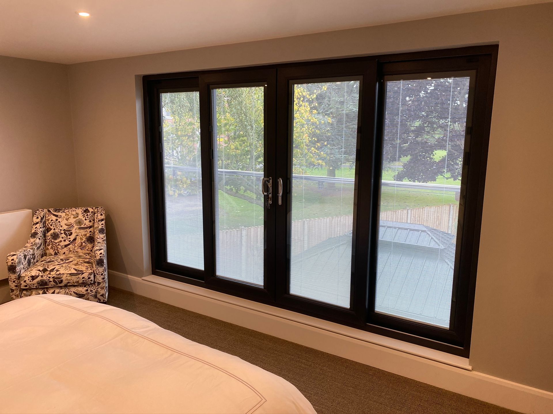 Bedroom with dark-framed sliding glass doors overlooking a green yard. A patterned armchair sits near the doors.