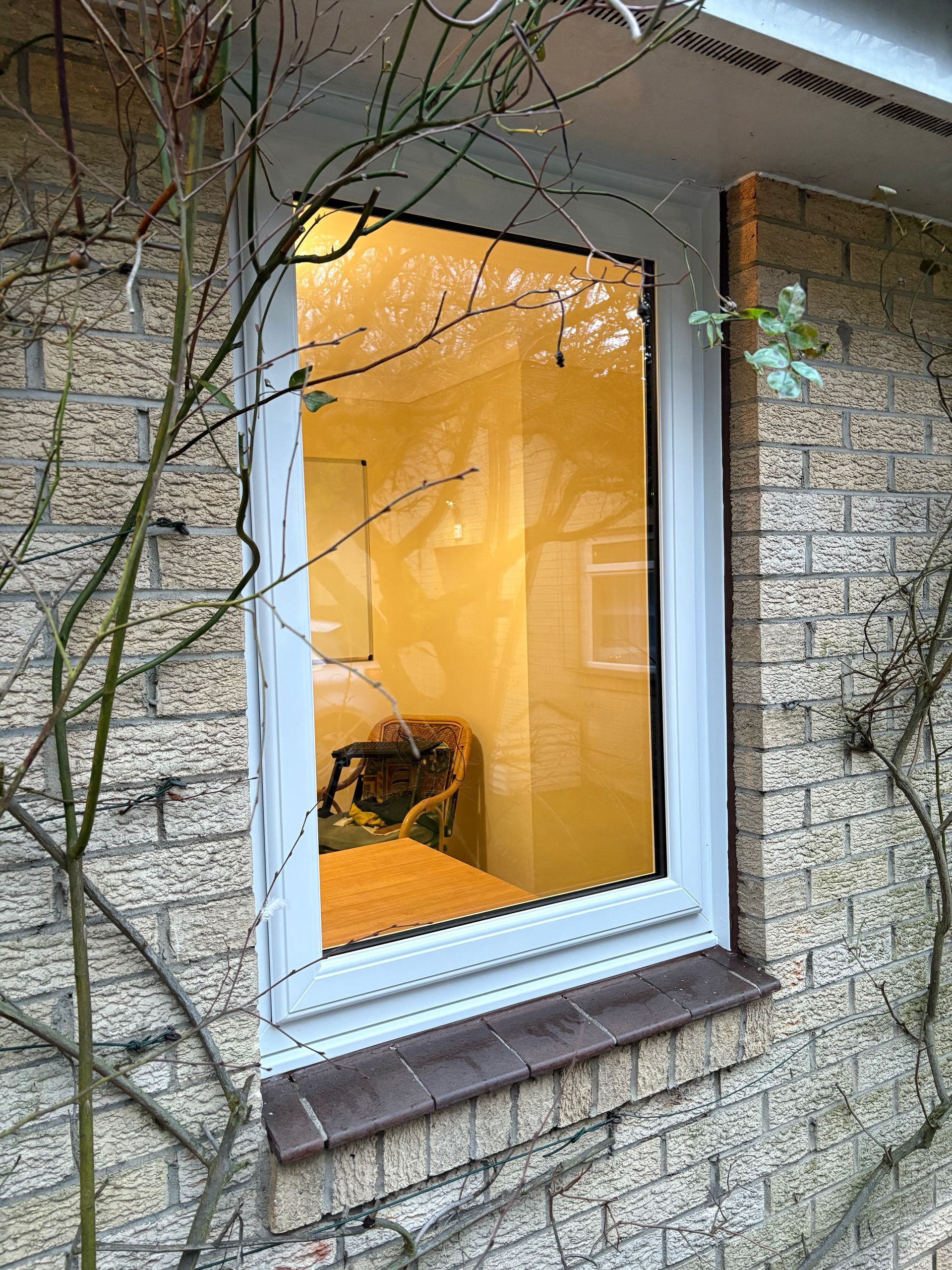 Window showing a room interior with warm yellow lighting, a table, and a chair, set against a brick wall exterior.