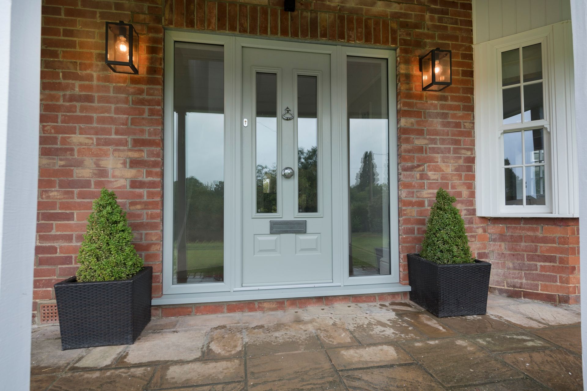 Gray front door with sidelights on a brick building, flanked by potted evergreens and sconce lights.