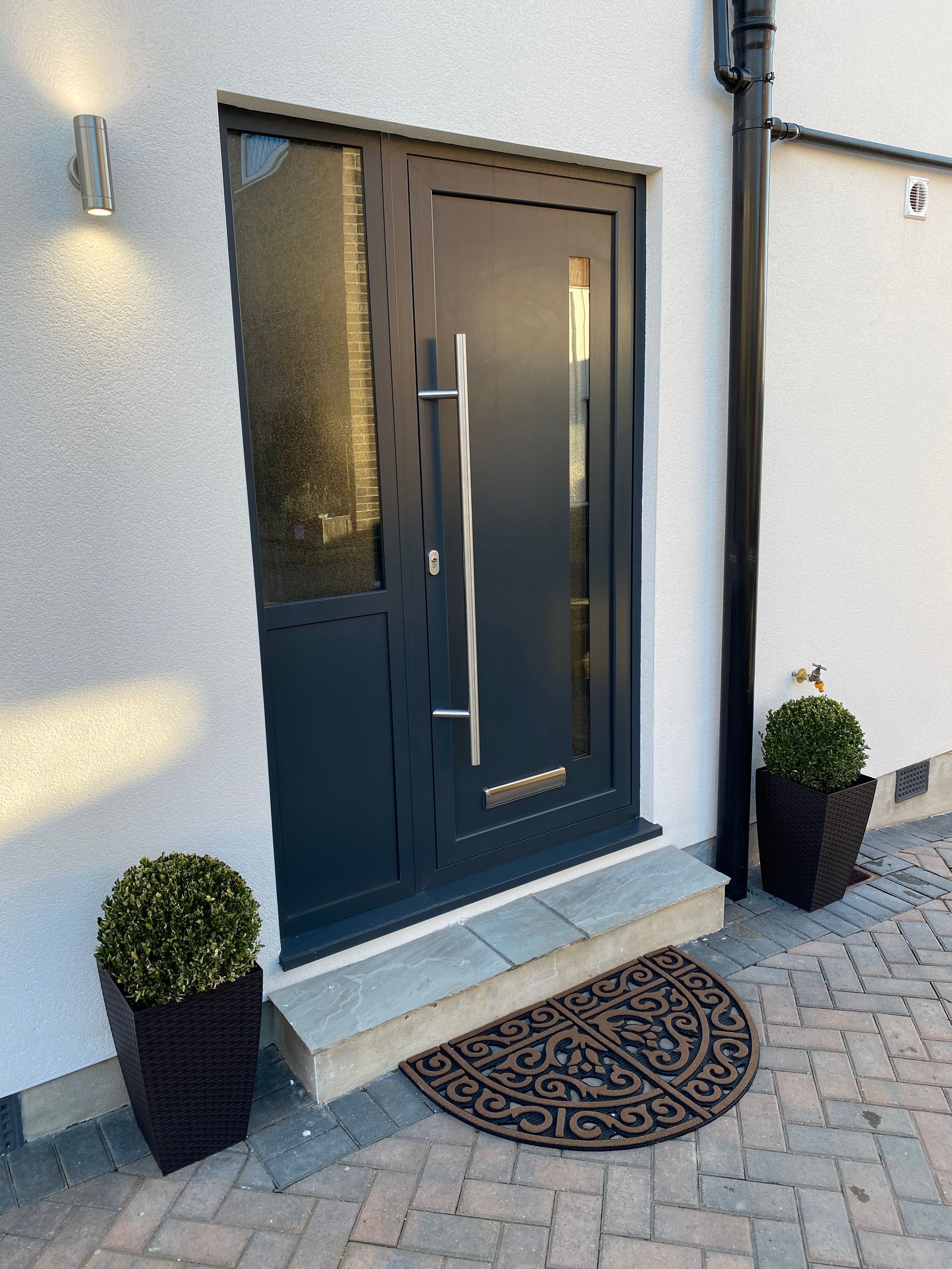Gray front door with sidelight, modern handle, welcome mat, and two potted plants, on a brick and stone surface.