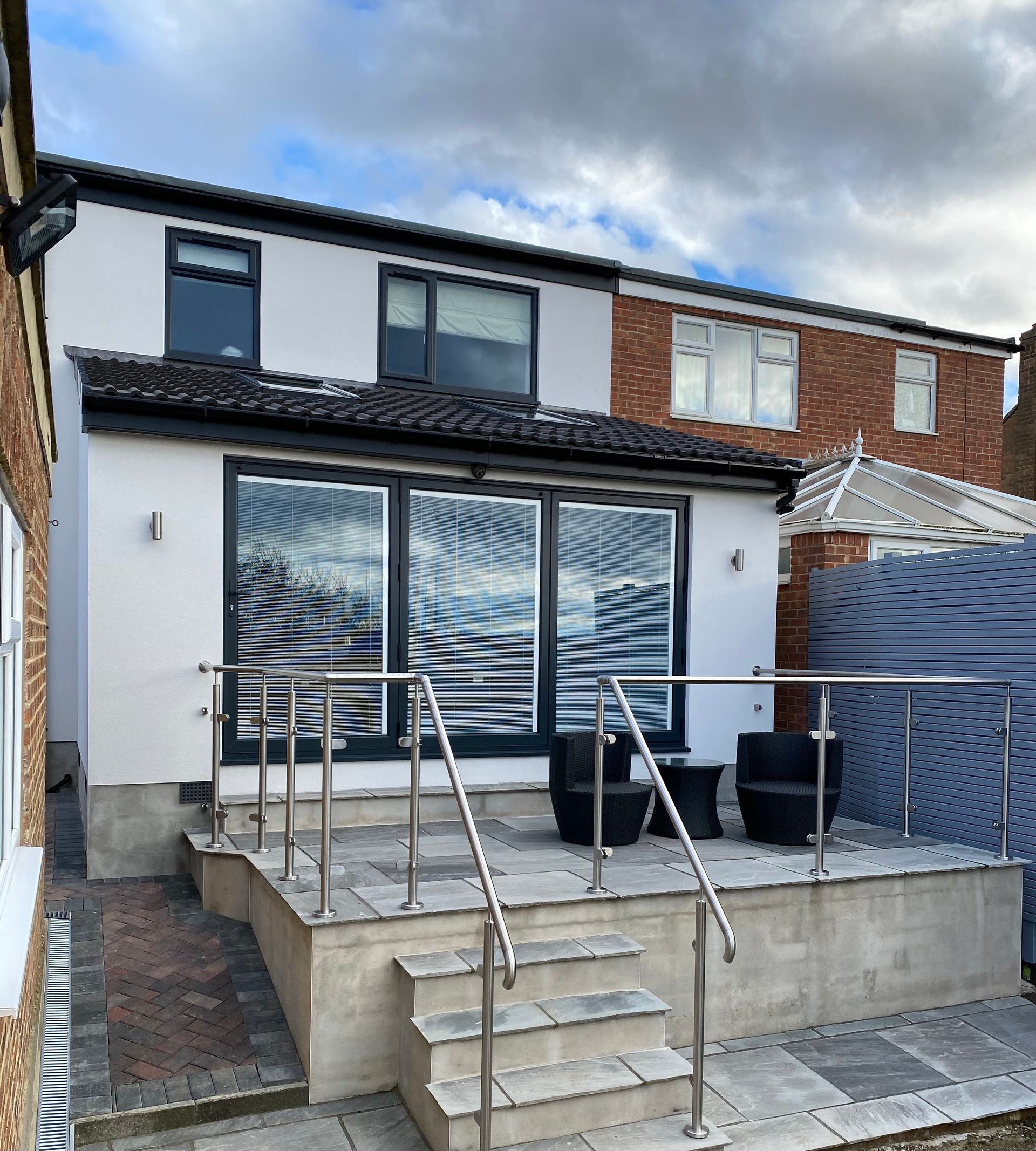 Back of a modern house with white walls, dark window frames, and a raised patio area with stainless steel railings and steps.