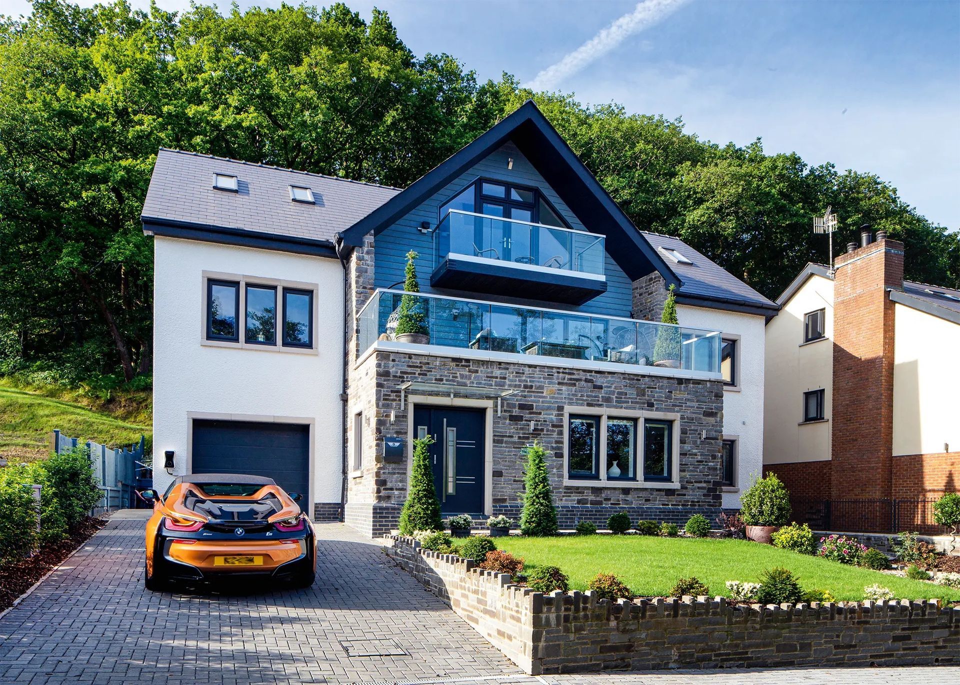 Modern two-story house with a luxury car in the driveway. The home has a stone facade, blue accents, and a balcony.