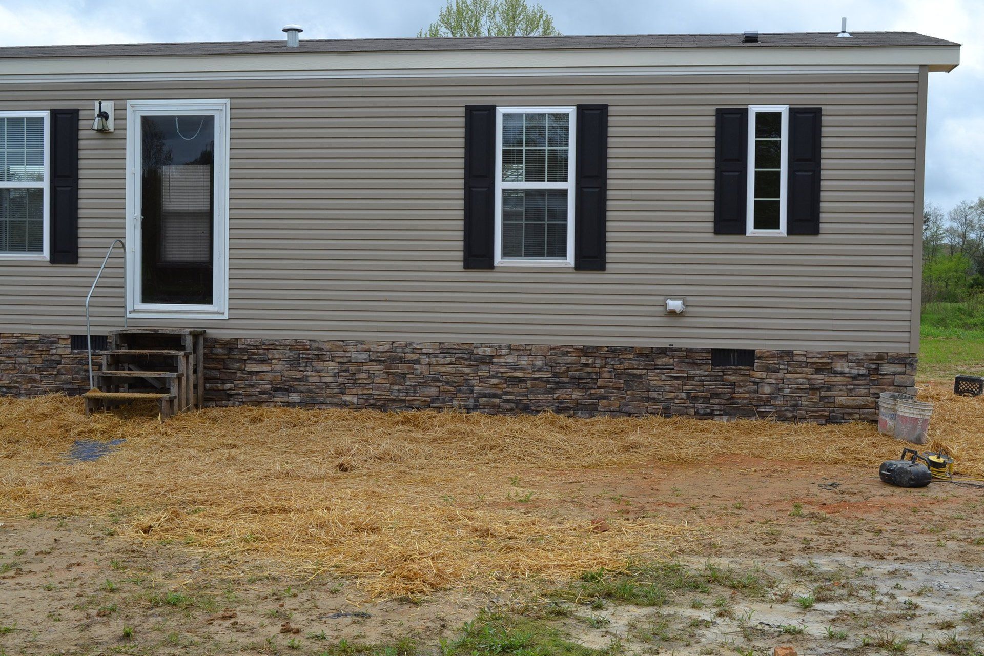 A mobile home with a stone siding and black shutters is sitting on top of a dirt field.