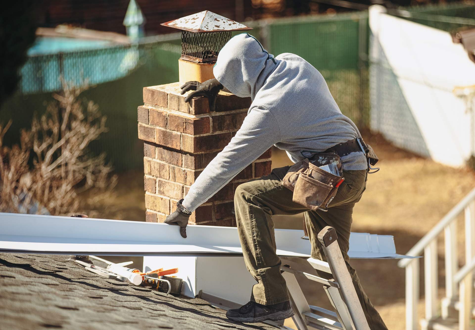 Worker in gray hoodie repairing a roof beside a brick chimney on a sunny day