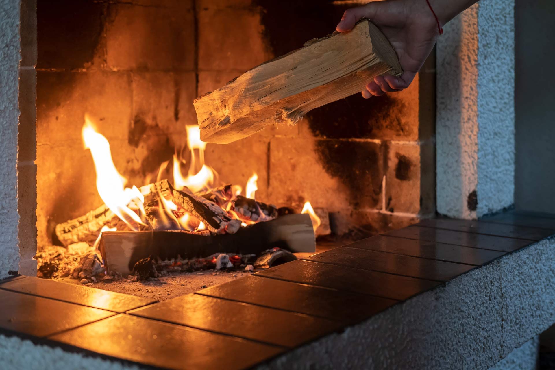 Hand placing a log into a burning fireplace with bright flames and glowing embers