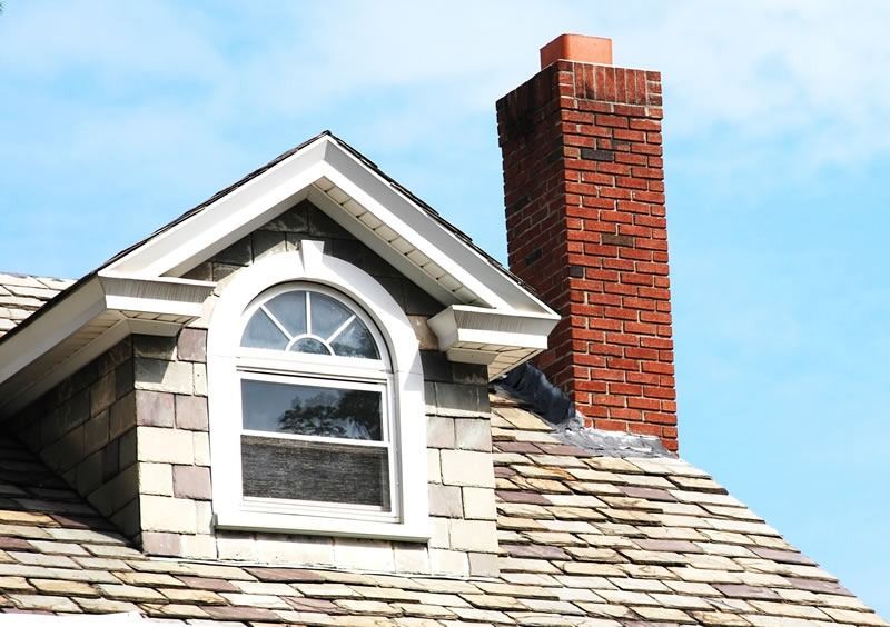 Dormer window and red brick chimney on a shingled roof under a blue sky