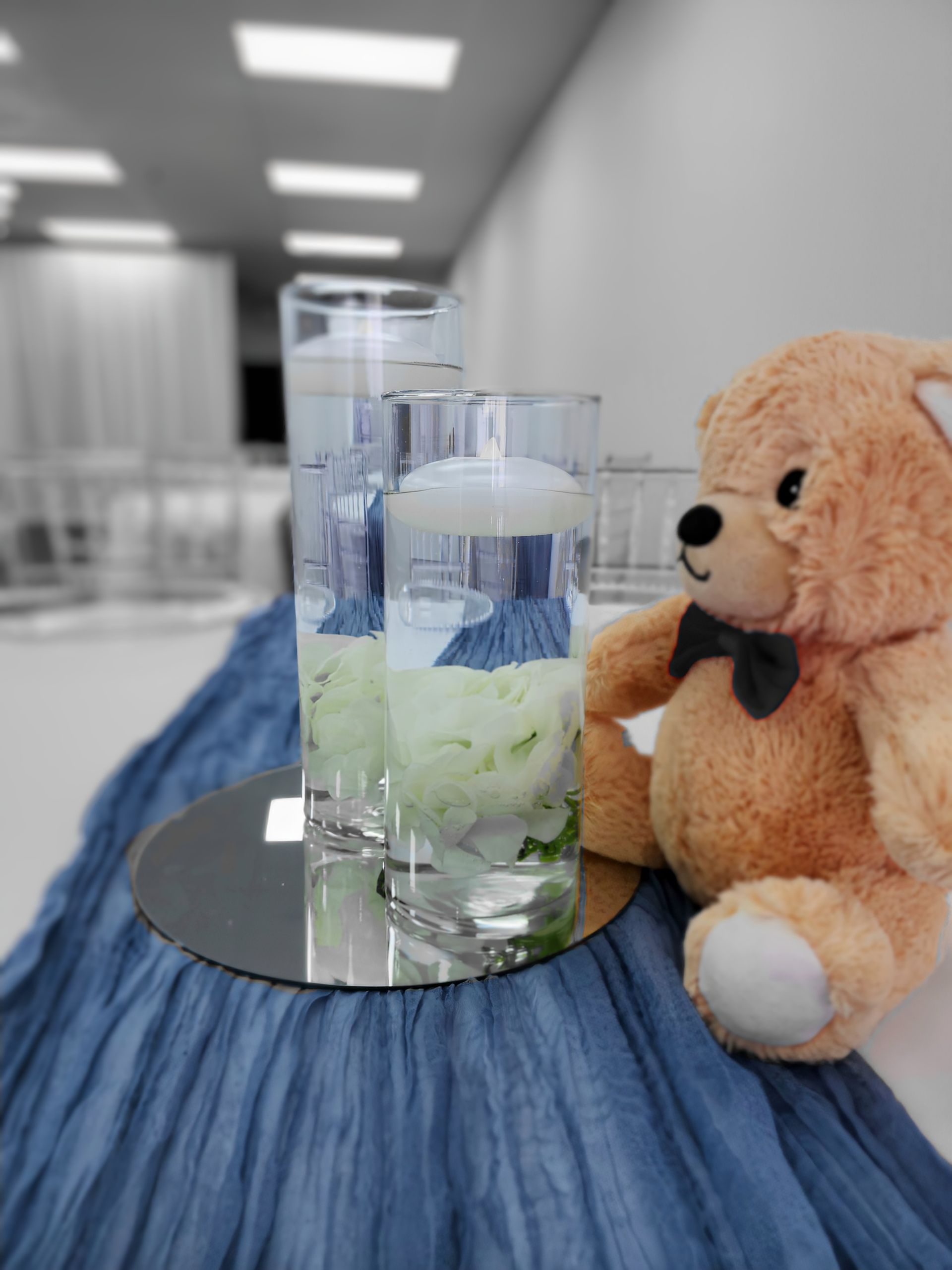 Teddy bear and floral centerpieces on blue cloth table.