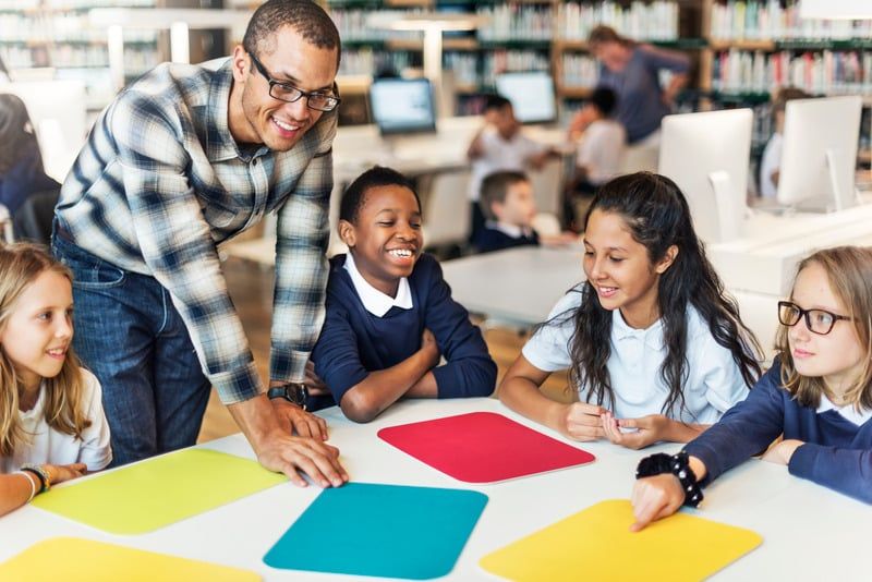 Teacher with a group of smiling students around a table with colorful squares in a library setting.