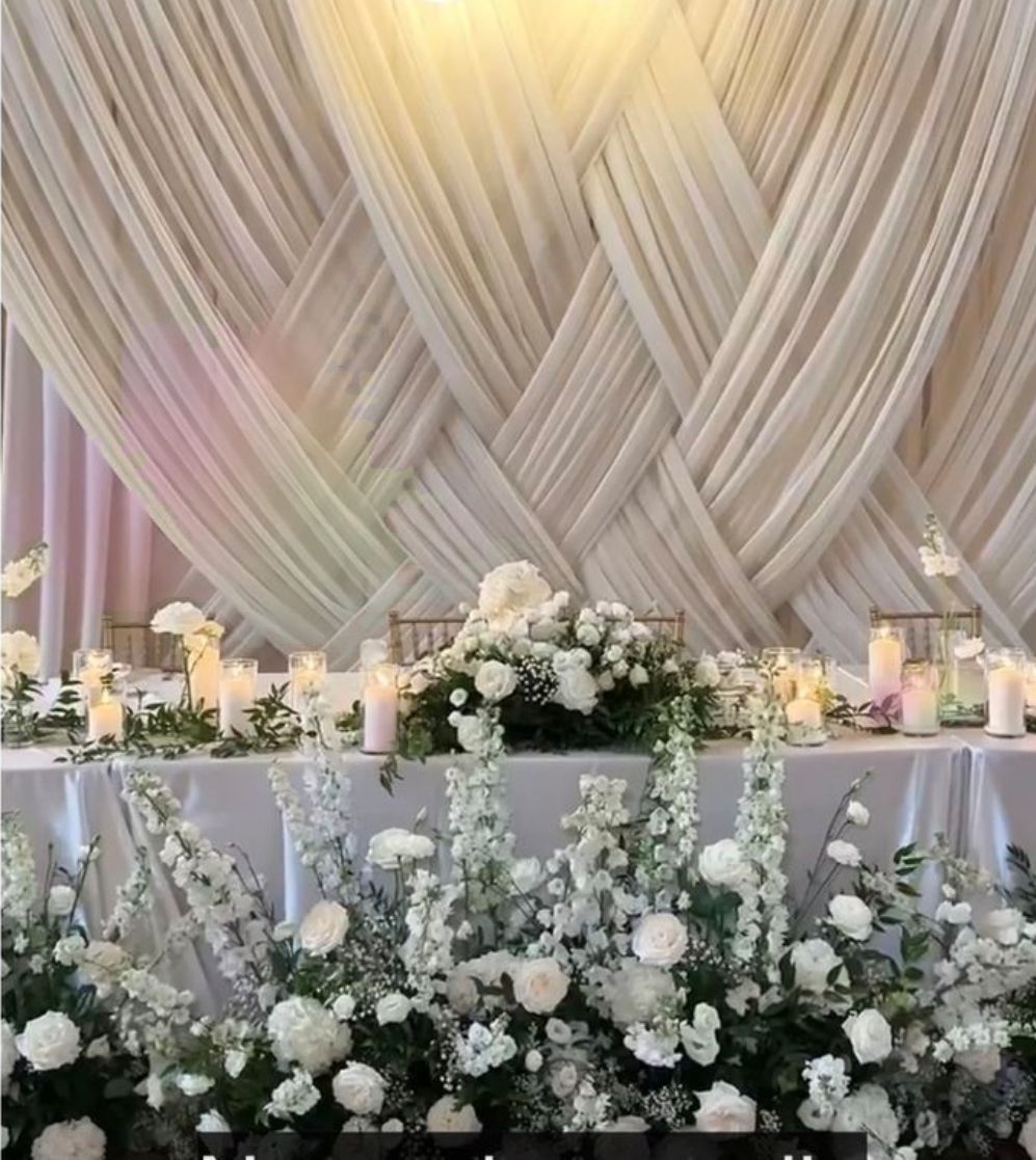 White wedding table decorated with flowers and candles, draped fabric backdrop.