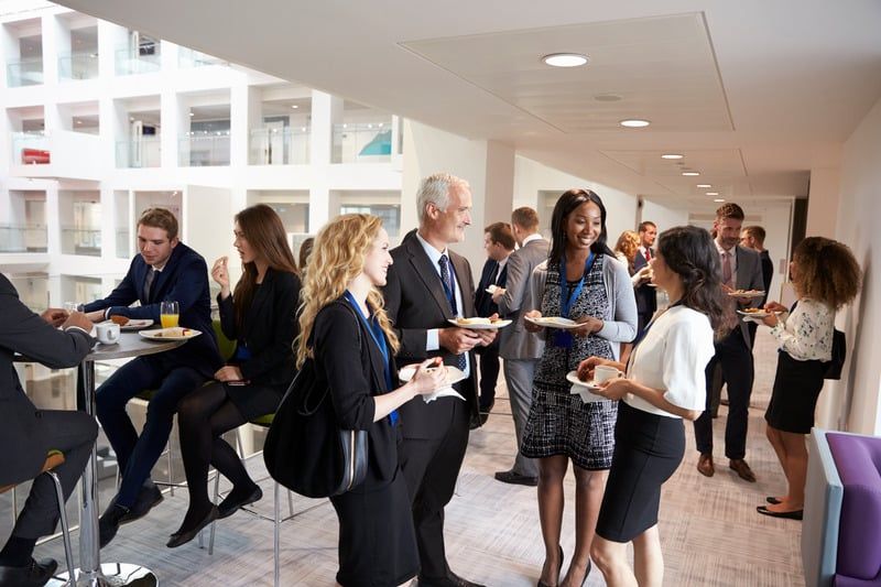 People in business attire socialize at a networking event in a modern, light-filled hallway, holding food and drinks.