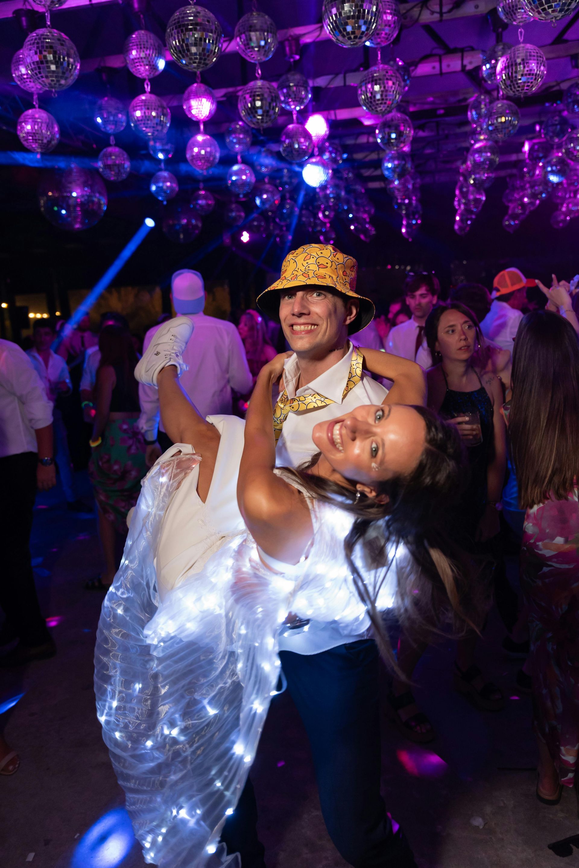 Two people dance; one carrying the other. Disco balls hang overhead. Vibrant purple and white lights.