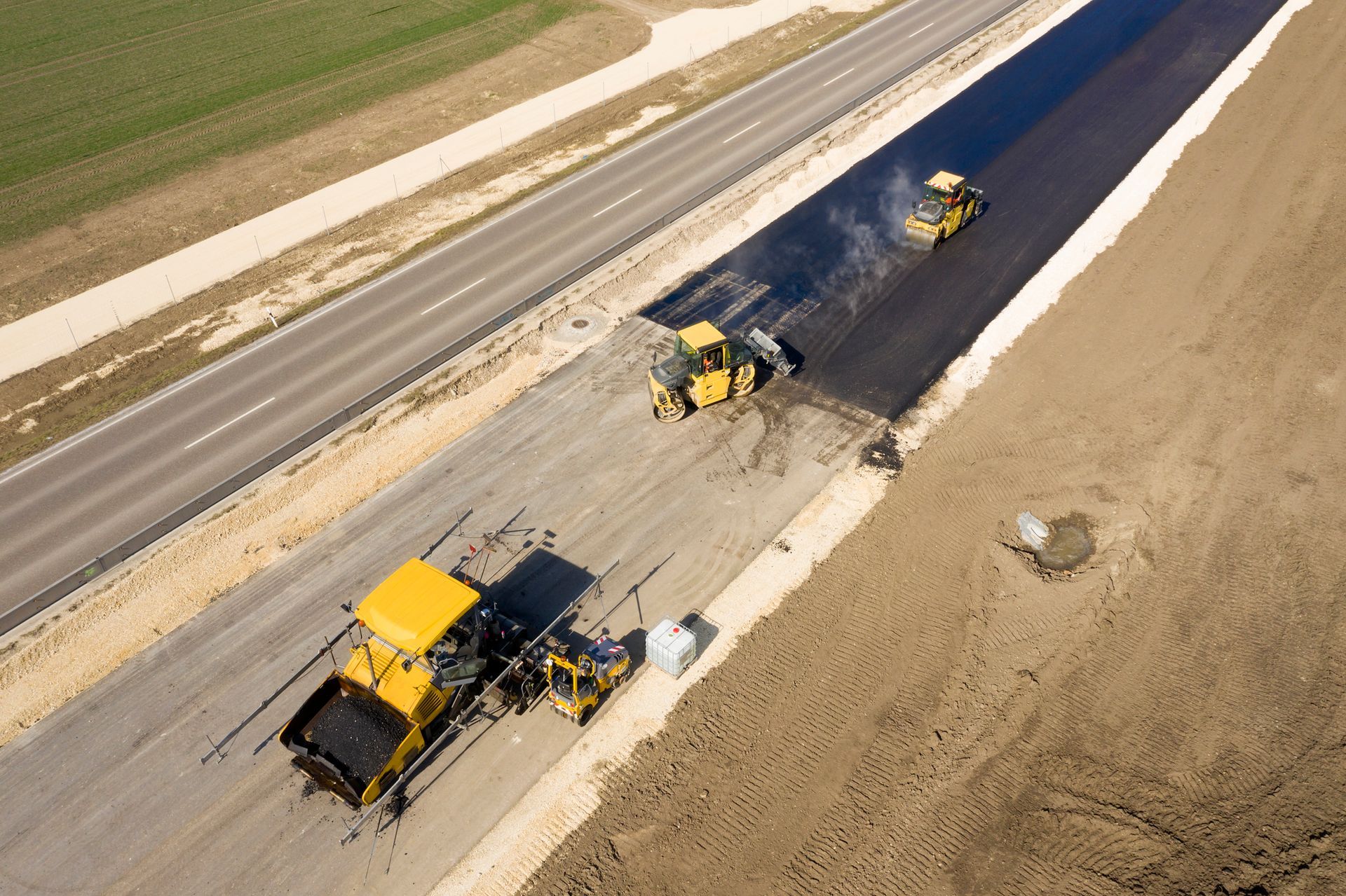 Road construction: Paving machines laying asphalt on new highway lane next to existing road, in a rural setting.