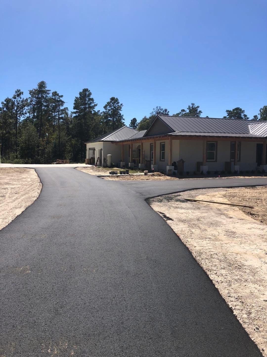 Paved driveway leading to a light-colored building under a clear, blue sky; surrounded by dirt and trees.