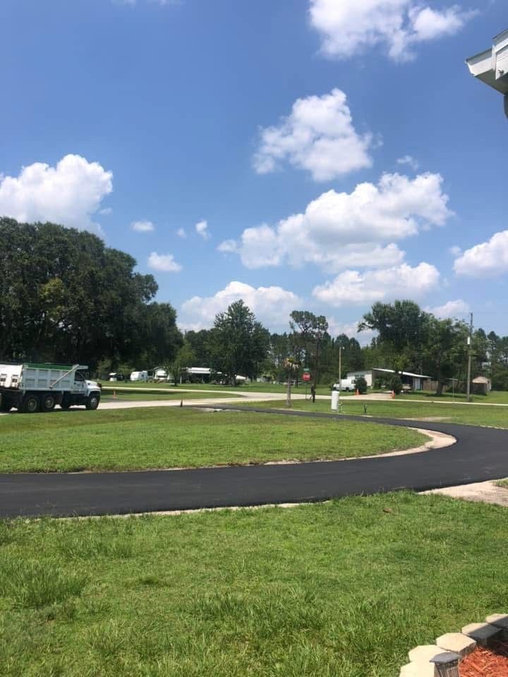 Road winding through a grassy area, with trees and houses under a partly cloudy blue sky.