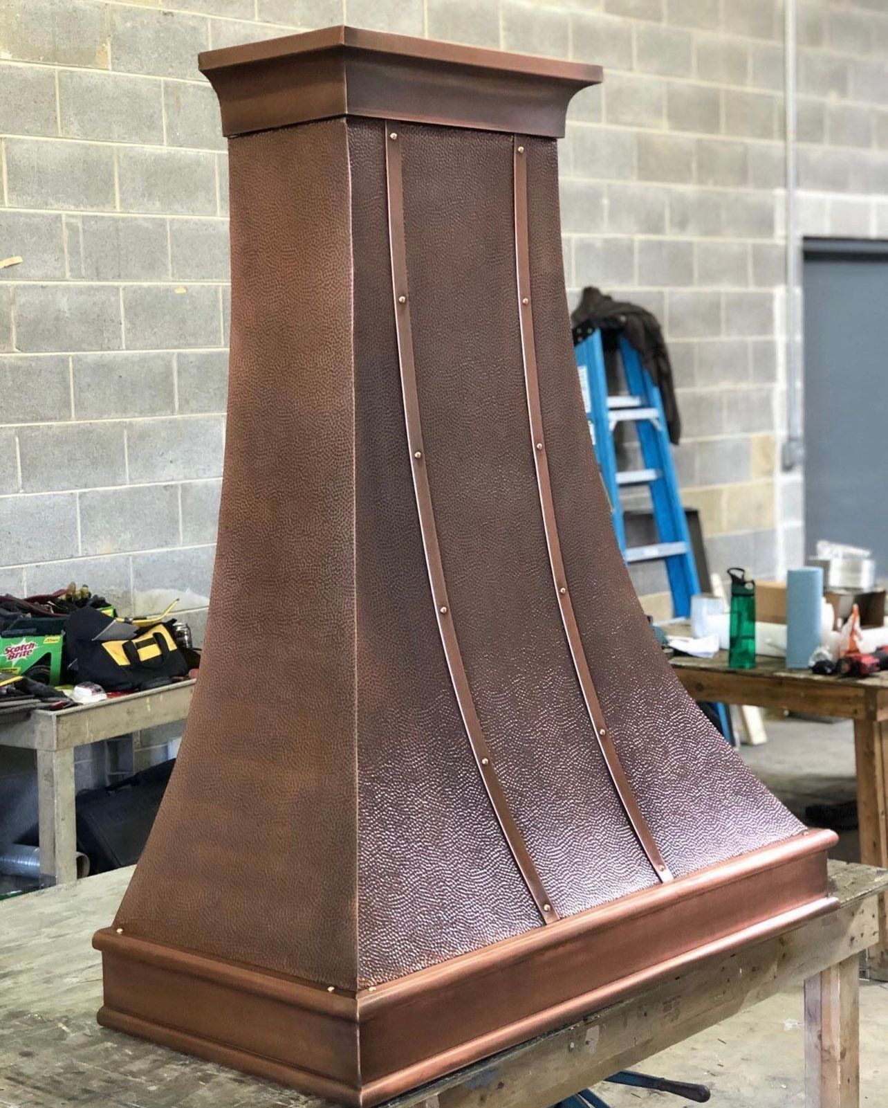 A copper hood is sitting on a wooden table in front of a brick wall.