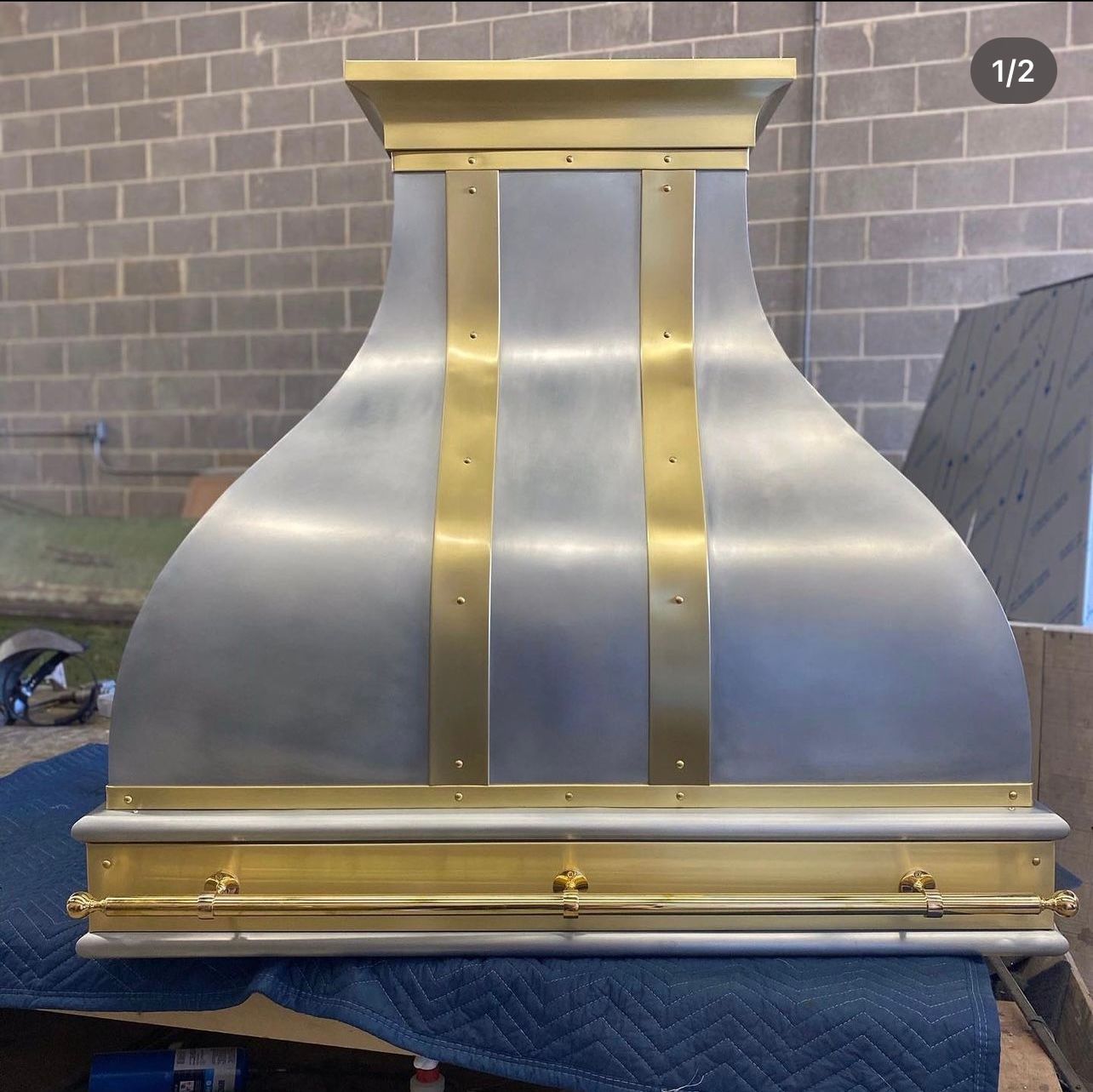 A stainless steel hood with brass trim sits on a blue blanket in front of a brick wall