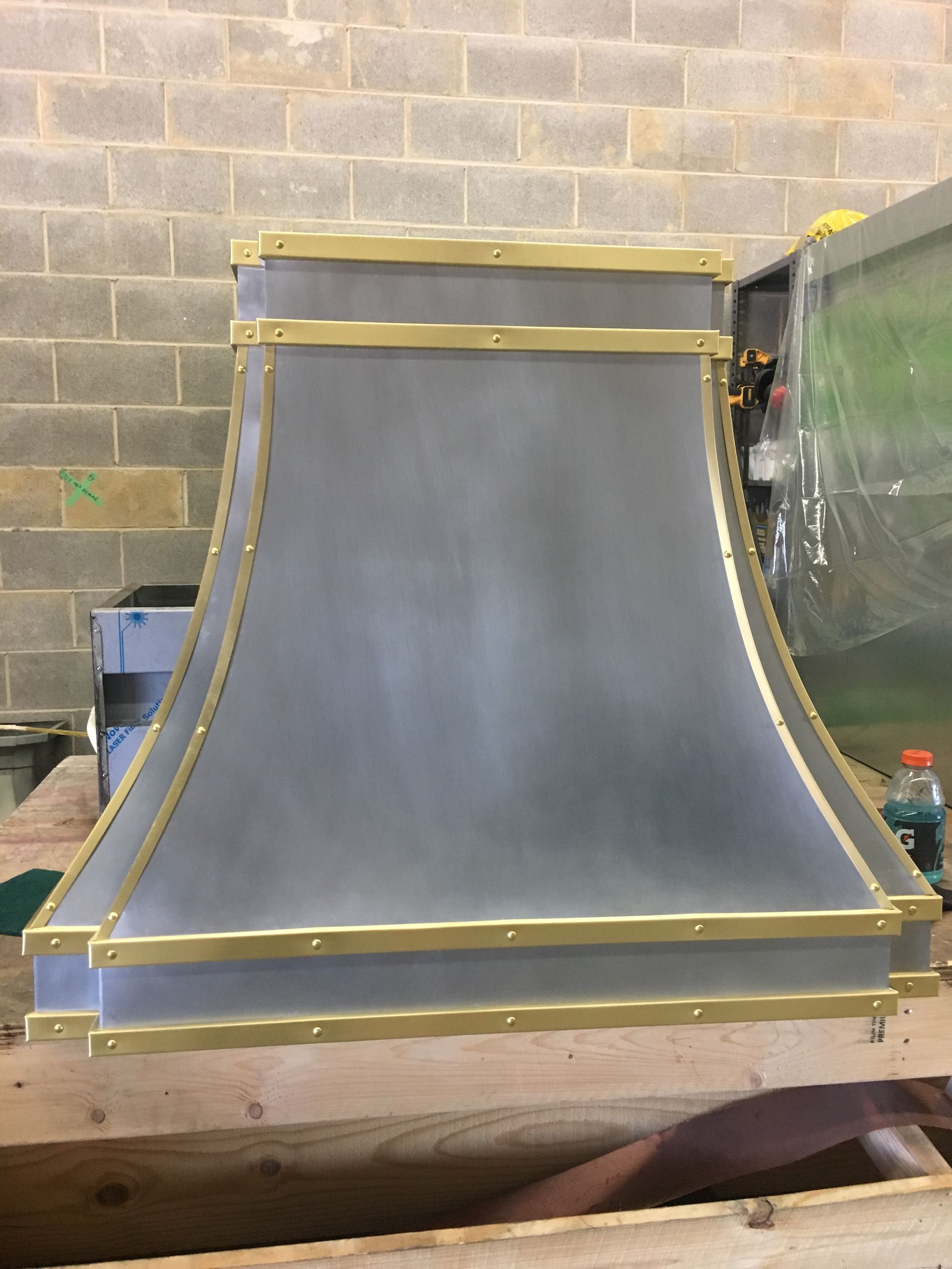 A metal hood with brass trim is sitting on a wooden table in front of a brick wall.