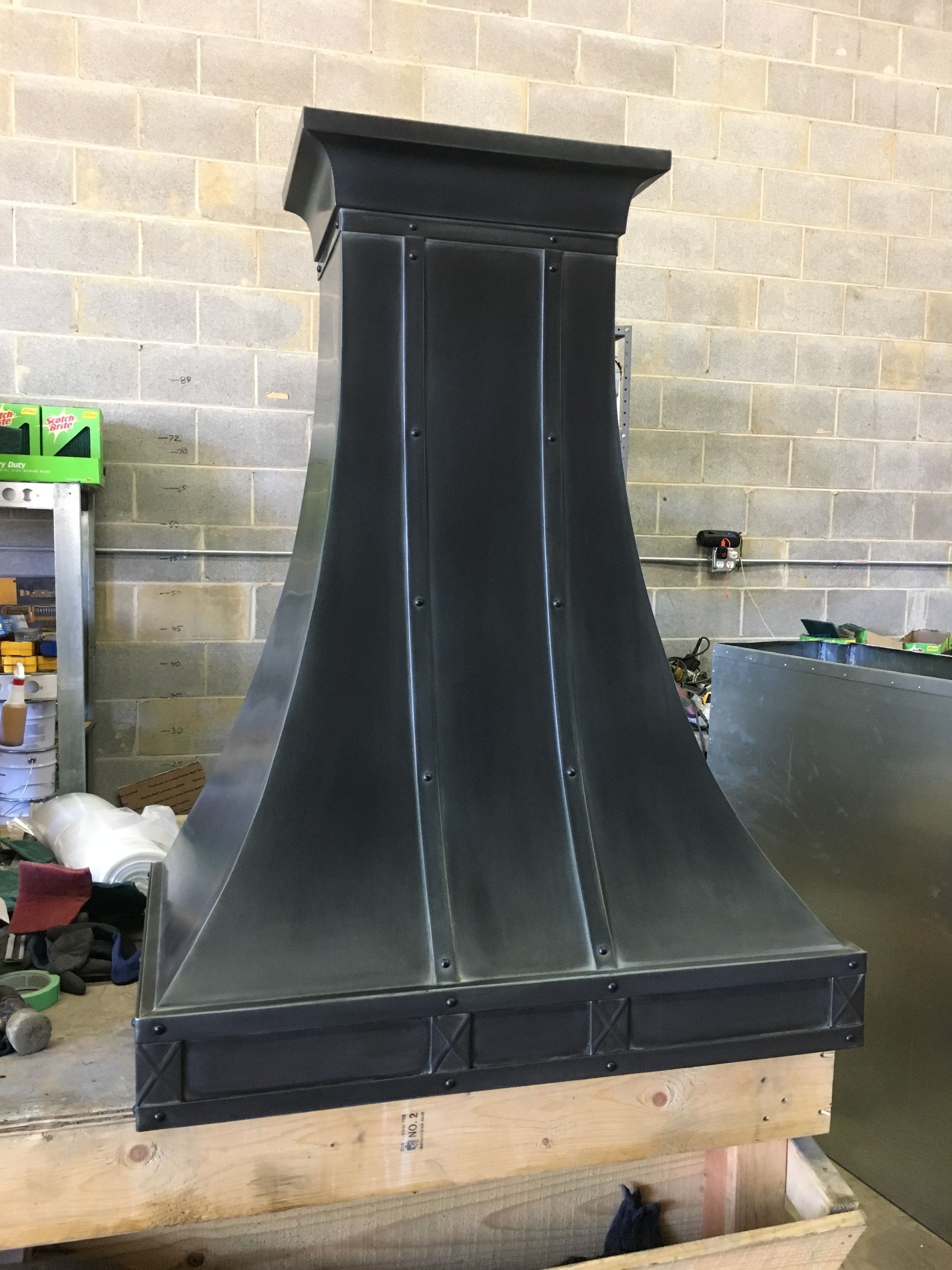 A black metal hood is sitting on a wooden table in front of a brick wall.