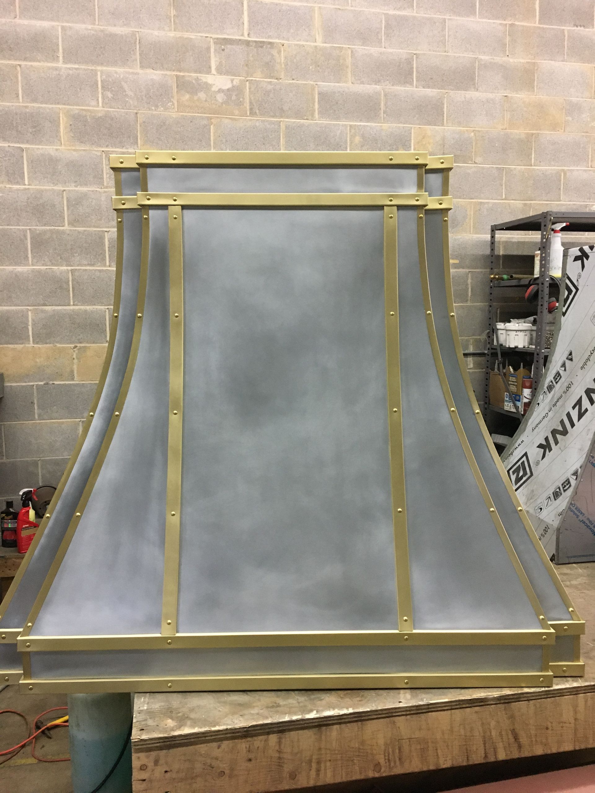 A metal hood with brass trim is sitting in front of a brick wall.