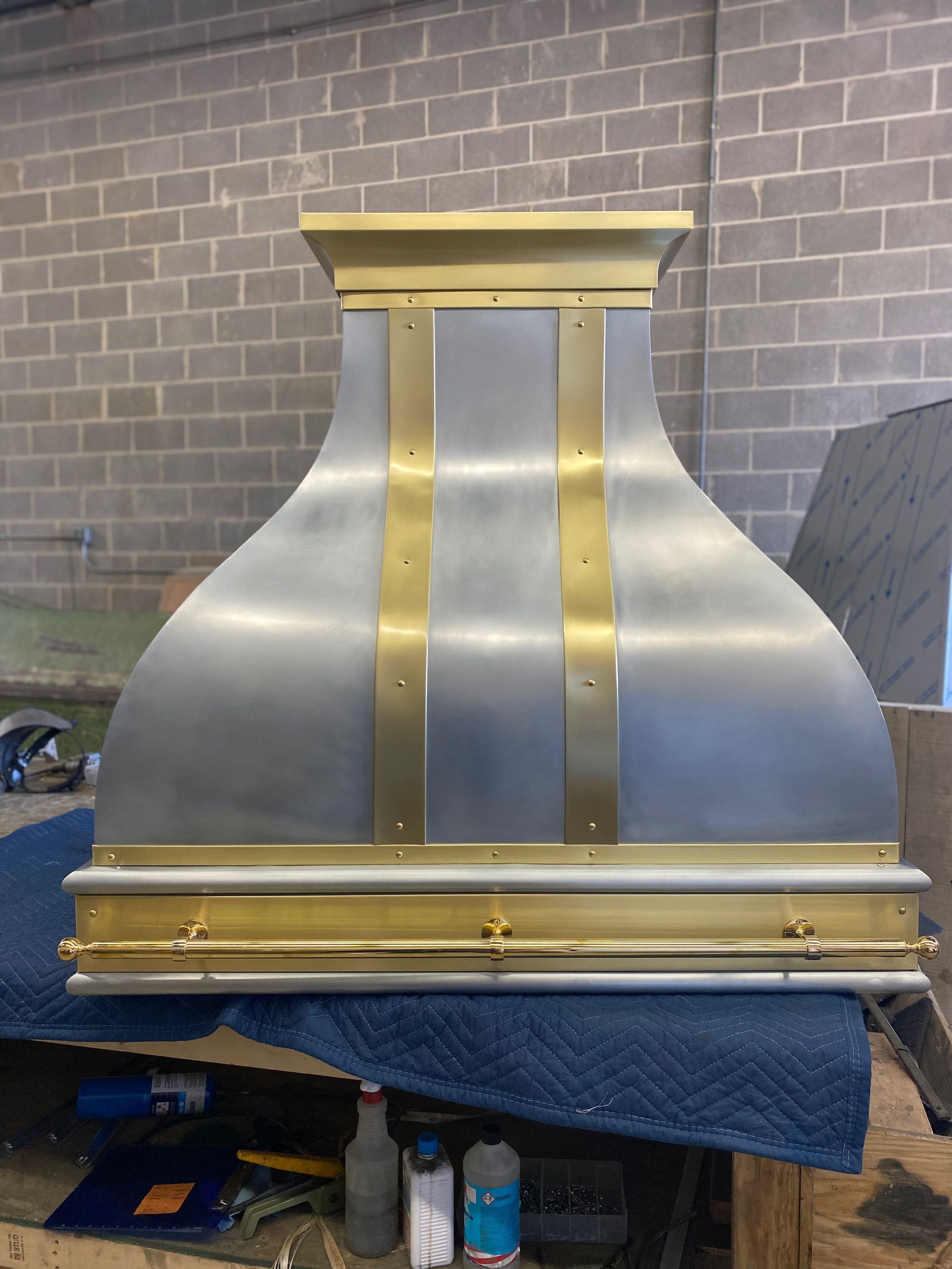 A stainless steel hood with brass trim is sitting on a table in front of a brick wall.