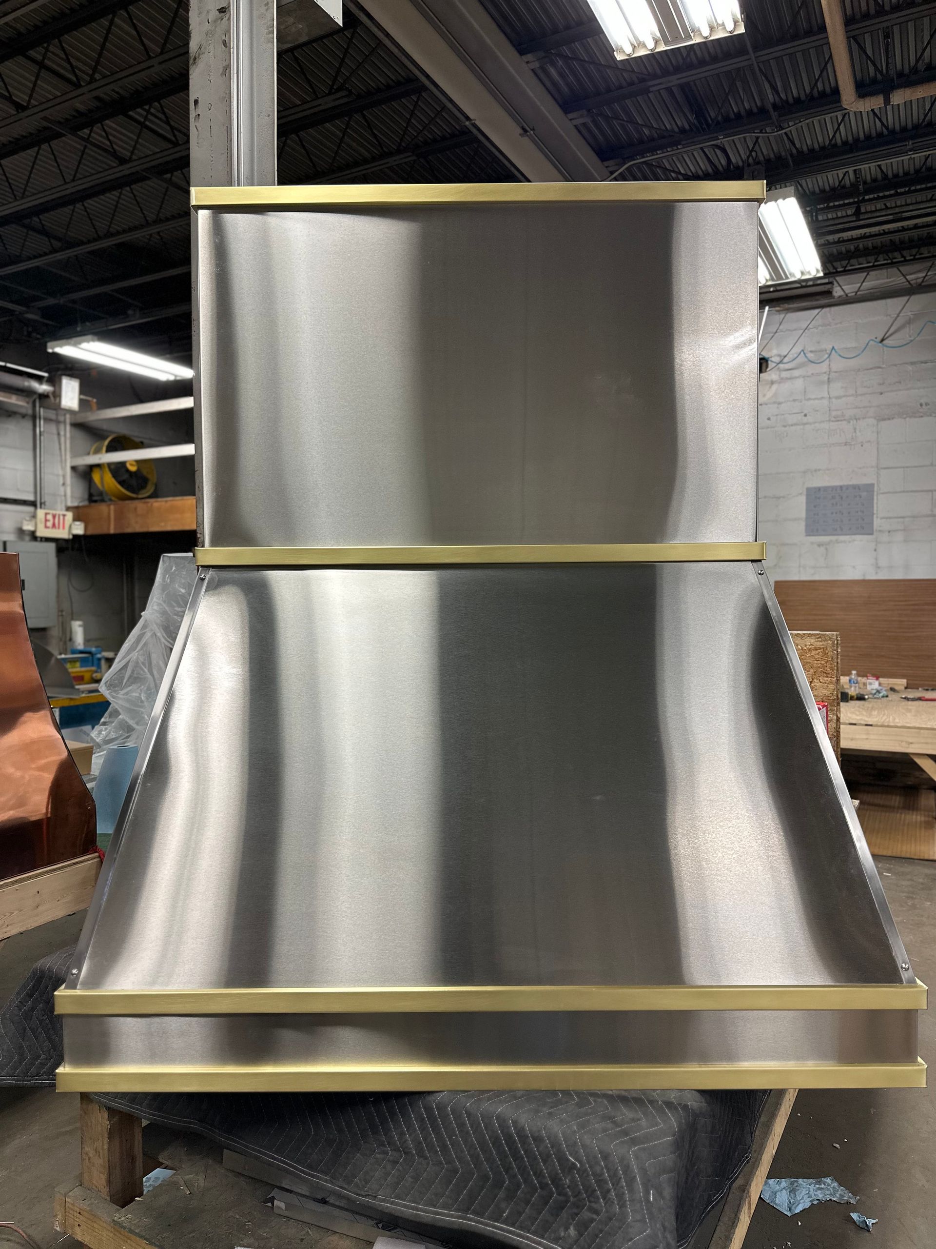 A stainless steel hood with brass trim is sitting on a table in a warehouse.