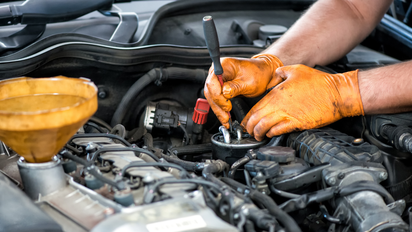 Mechanic wearing orange gloves repairs a car engine in the open hood area.
