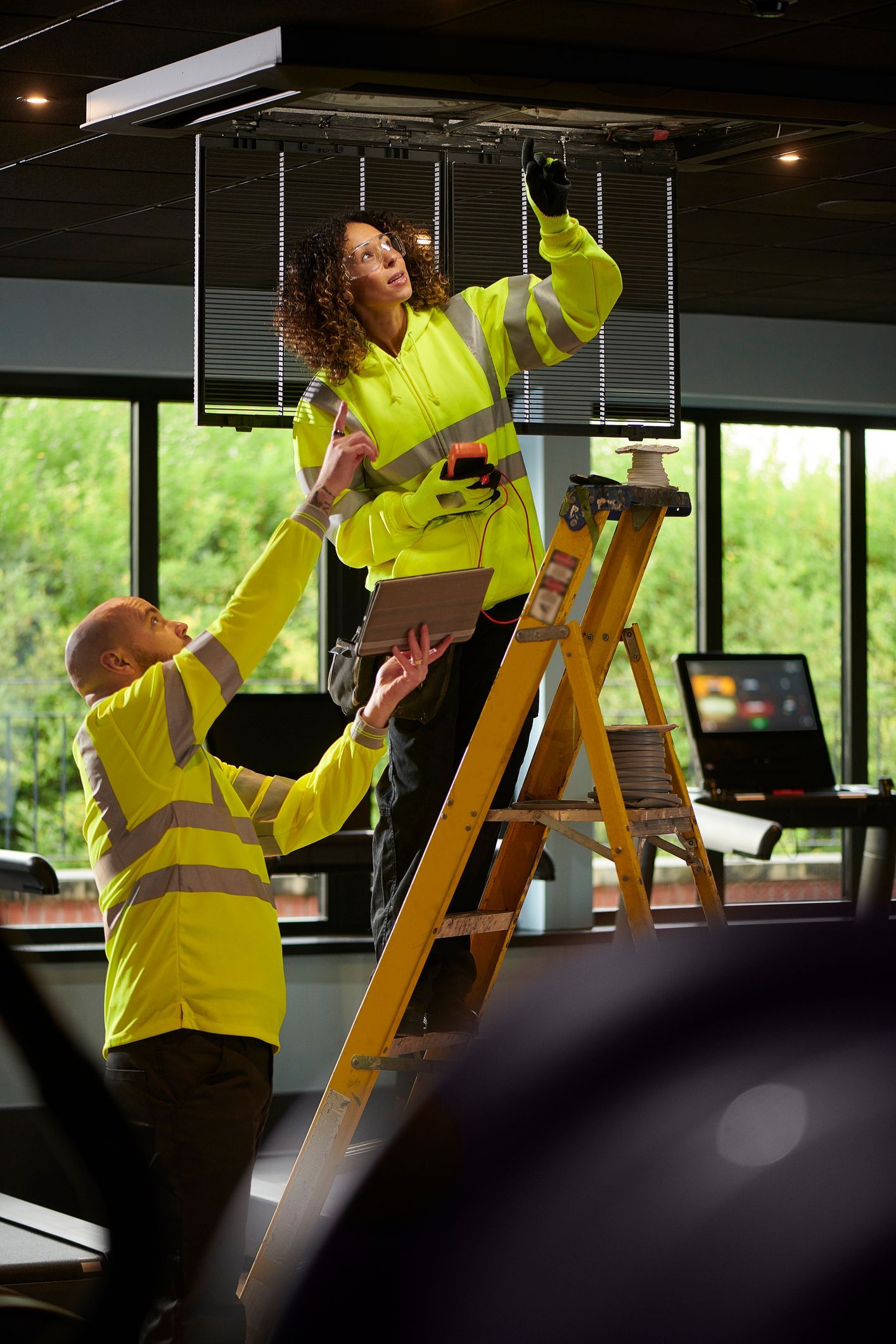 Two workers in safety vests, one on a ladder, adjusting overhead lighting