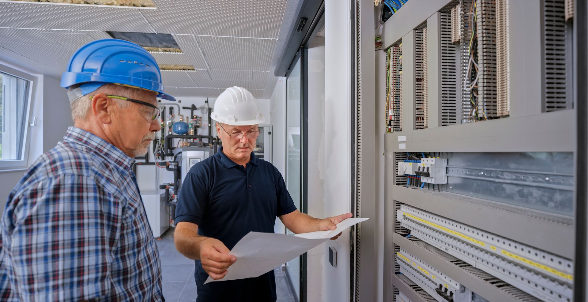 Two people in hard hats examine an electrical panel, one holding a blueprint