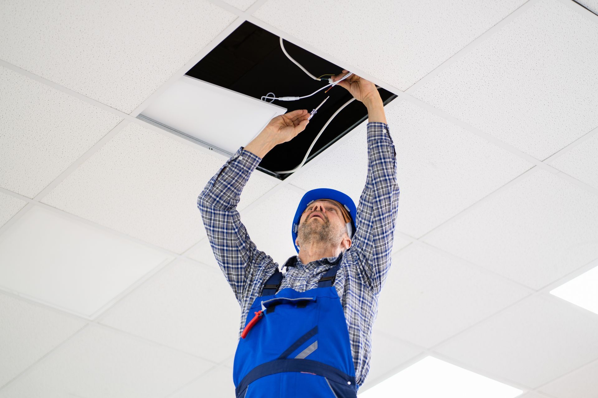 Electrician in blue overalls and a hard hat is working on the ceiling wiring