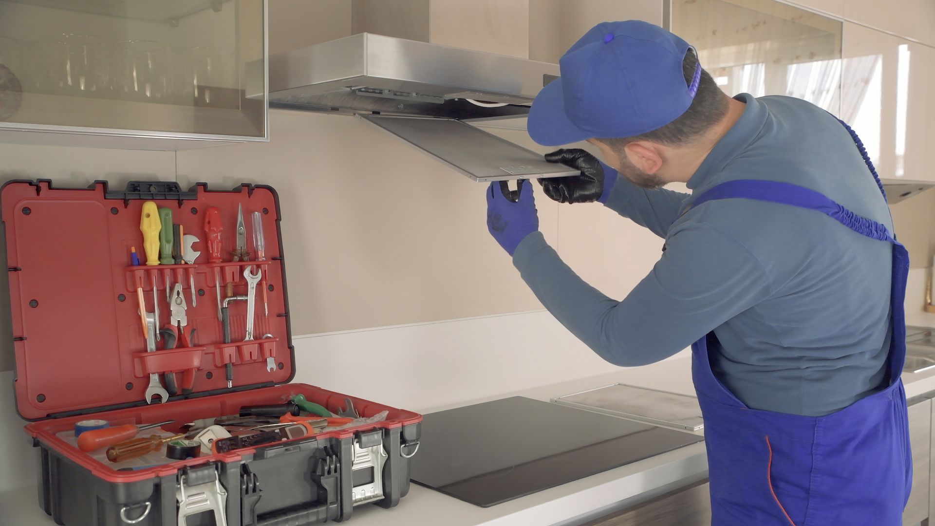 A repairman in blue overalls and a cap works on a kitchen range hood