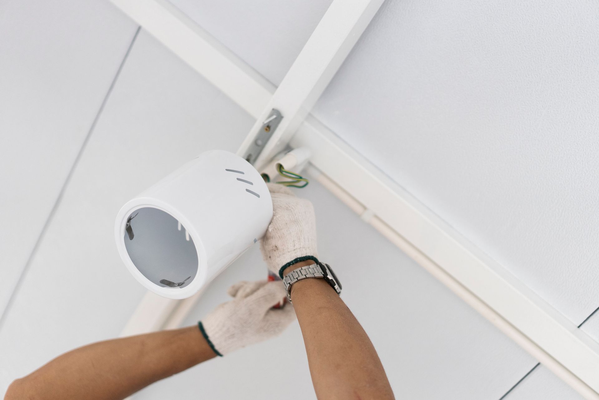 A person wearing gloves is installing a white cylindrical light fixture on a ceiling.