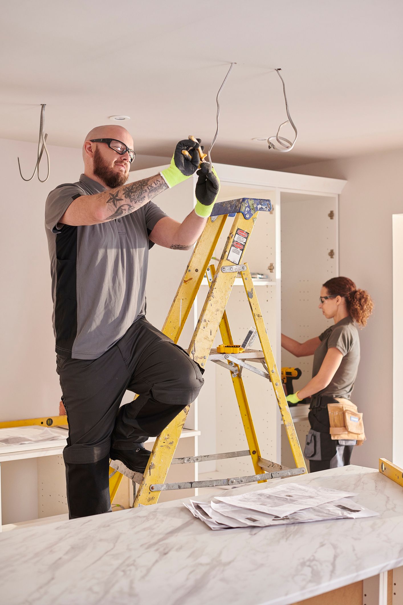 Two people are renovating a room. One person on a ladder works on the ceiling wires
