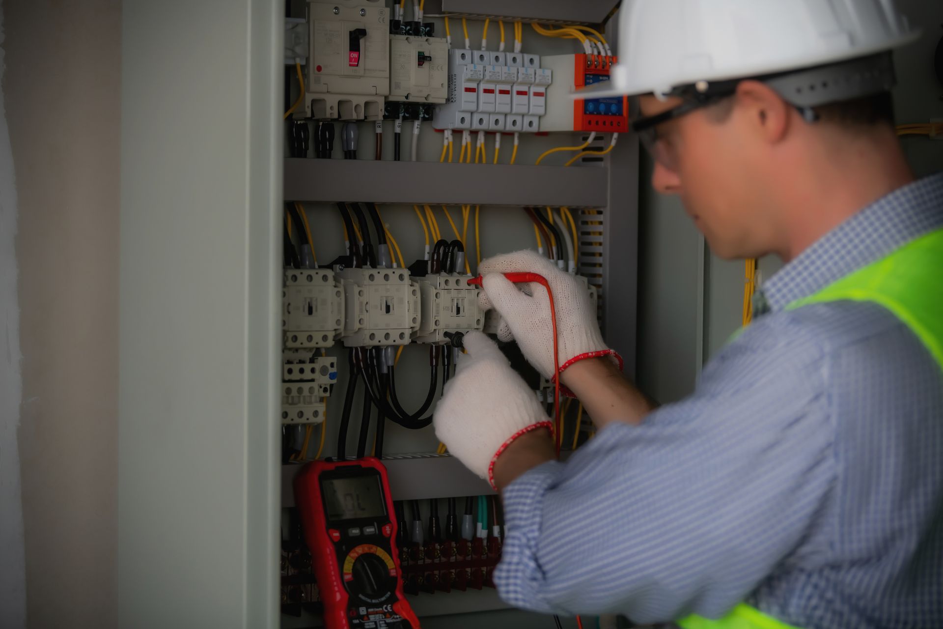Electrician in a safety vest and a hard hat using a multimeter on an electrical panel