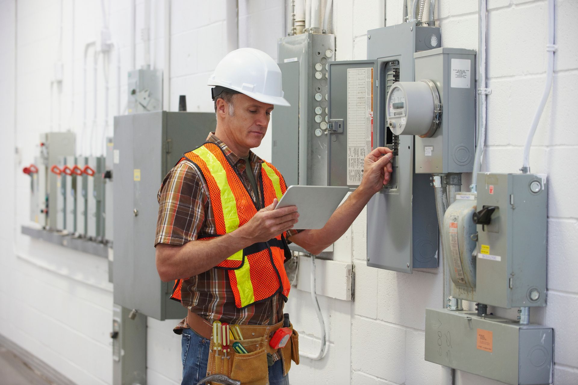 An electrician in a hard hat and vest inspects the electrical panel, using a tablet