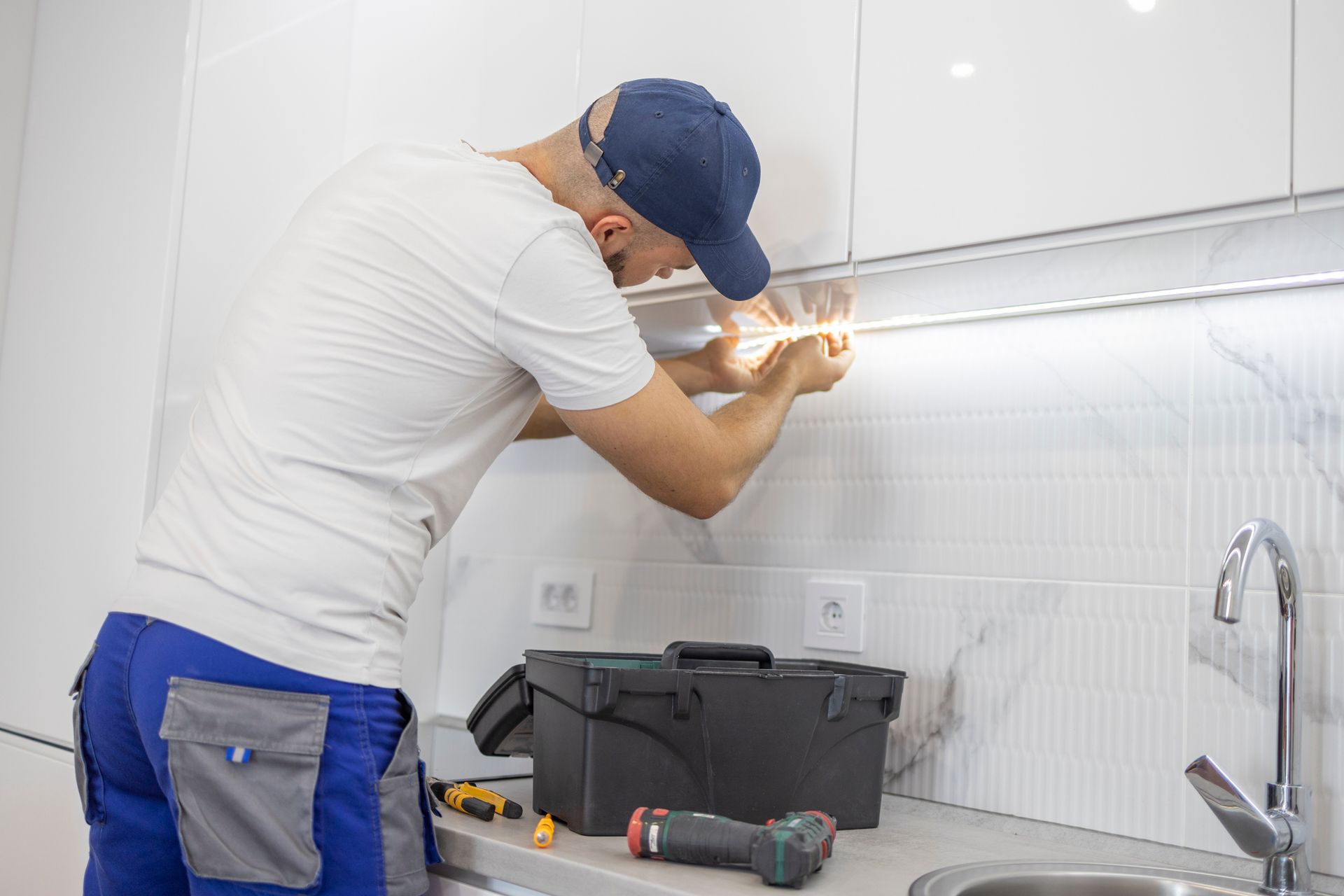 A person in work clothes is installing under-cabinet lighting in a kitchen