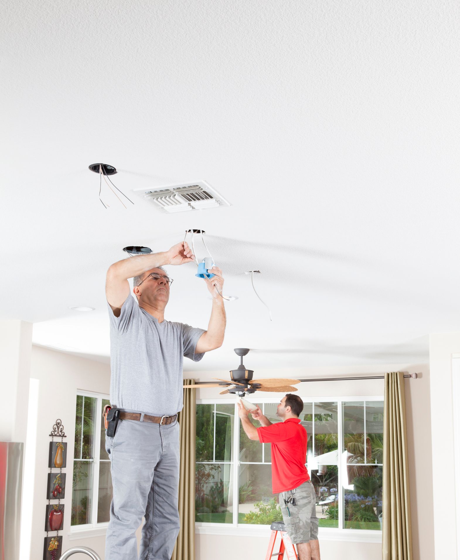 Two people are installing ceiling lights and a ceiling fan in a house