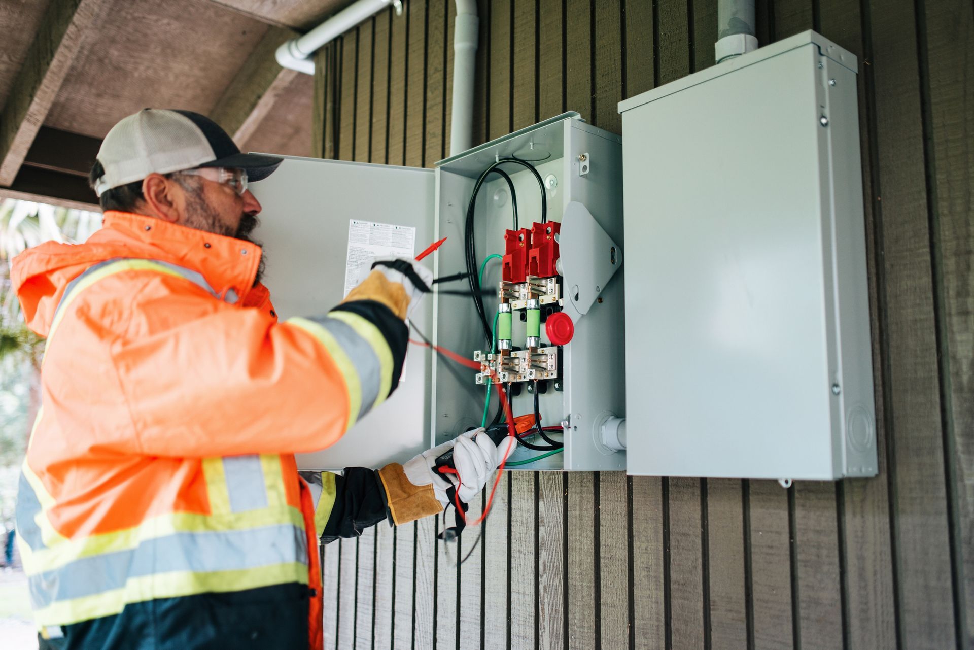 An electrician in an orange safety vest, working on an electrical box on an exterior building